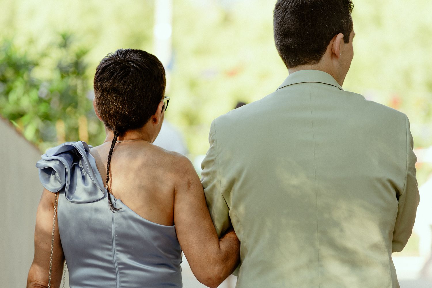 A couple walks together arm in arm, seen from behind, dressed in formal attire outdoors.
