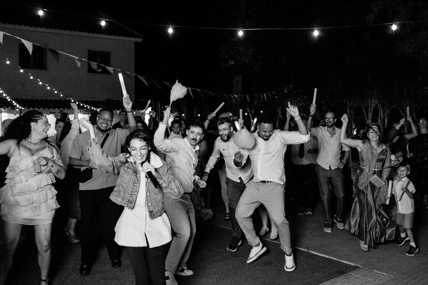 Black and white photo of joyful wedding guests dancing and celebrating outdoors at a festive night reception.