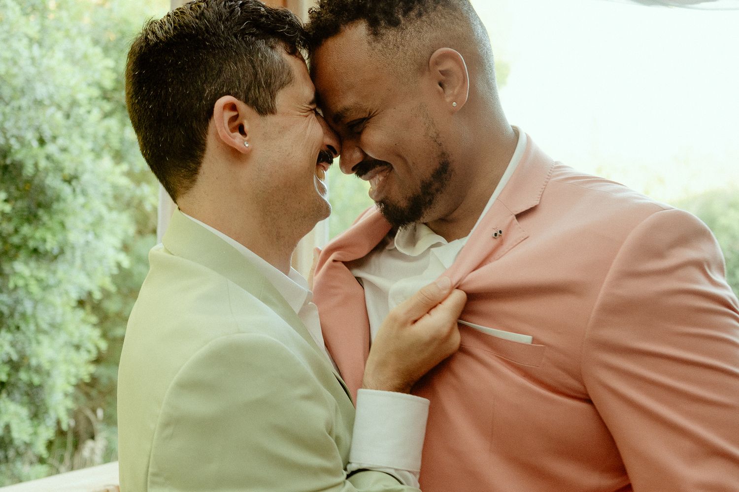 Two grooms in pastel suits share an intimate moment, touching foreheads and smiling at their wedding celebration.