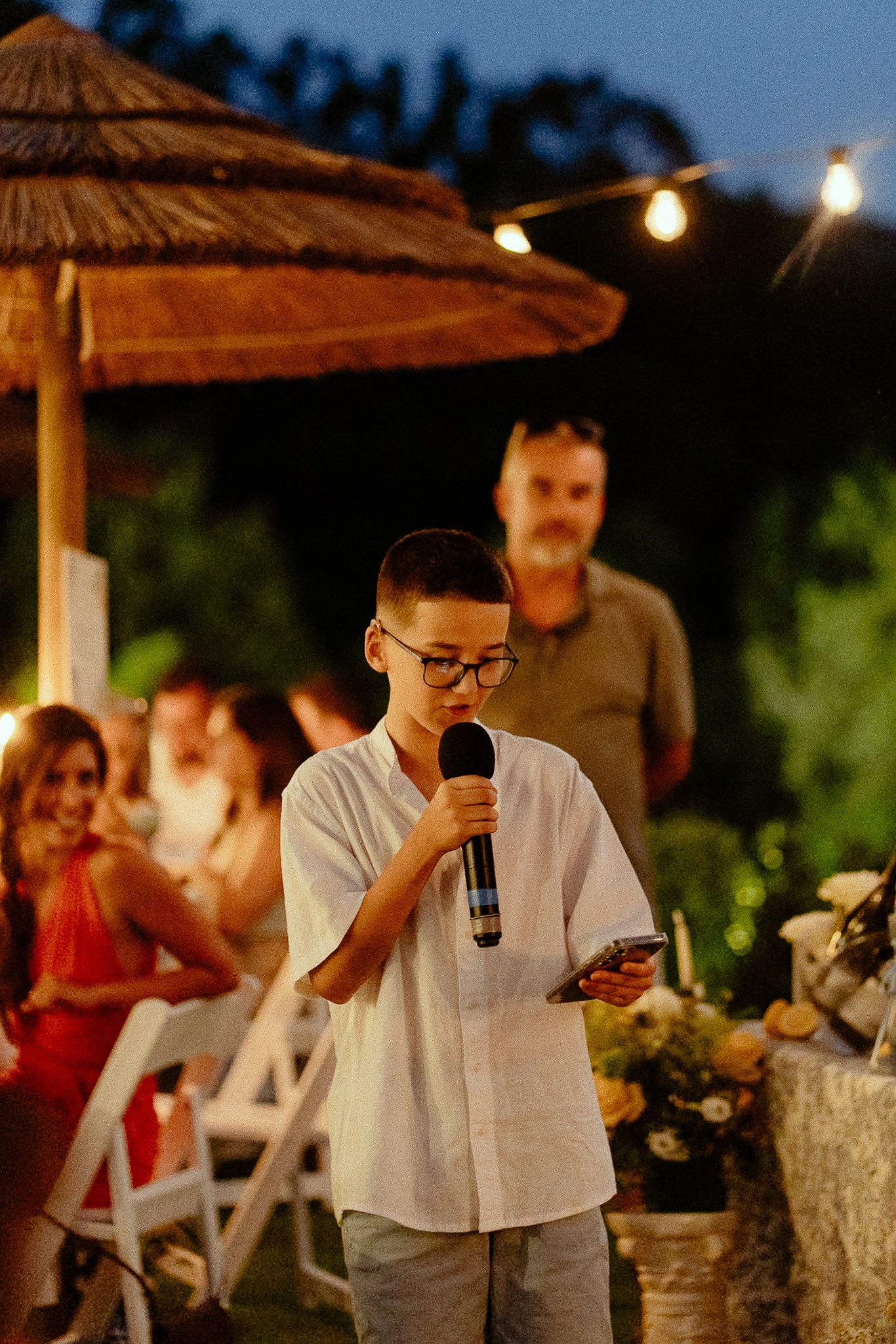 A young boy in glasses speaks into a microphone at an outdoor evening party with string lights.