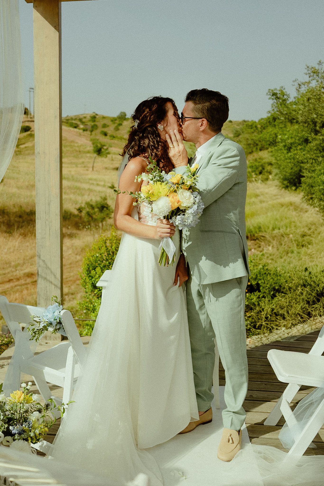 Bride and groom share first kiss at outdoor wedding ceremony, bride holding yellow and white bouquet, green hills backdrop.