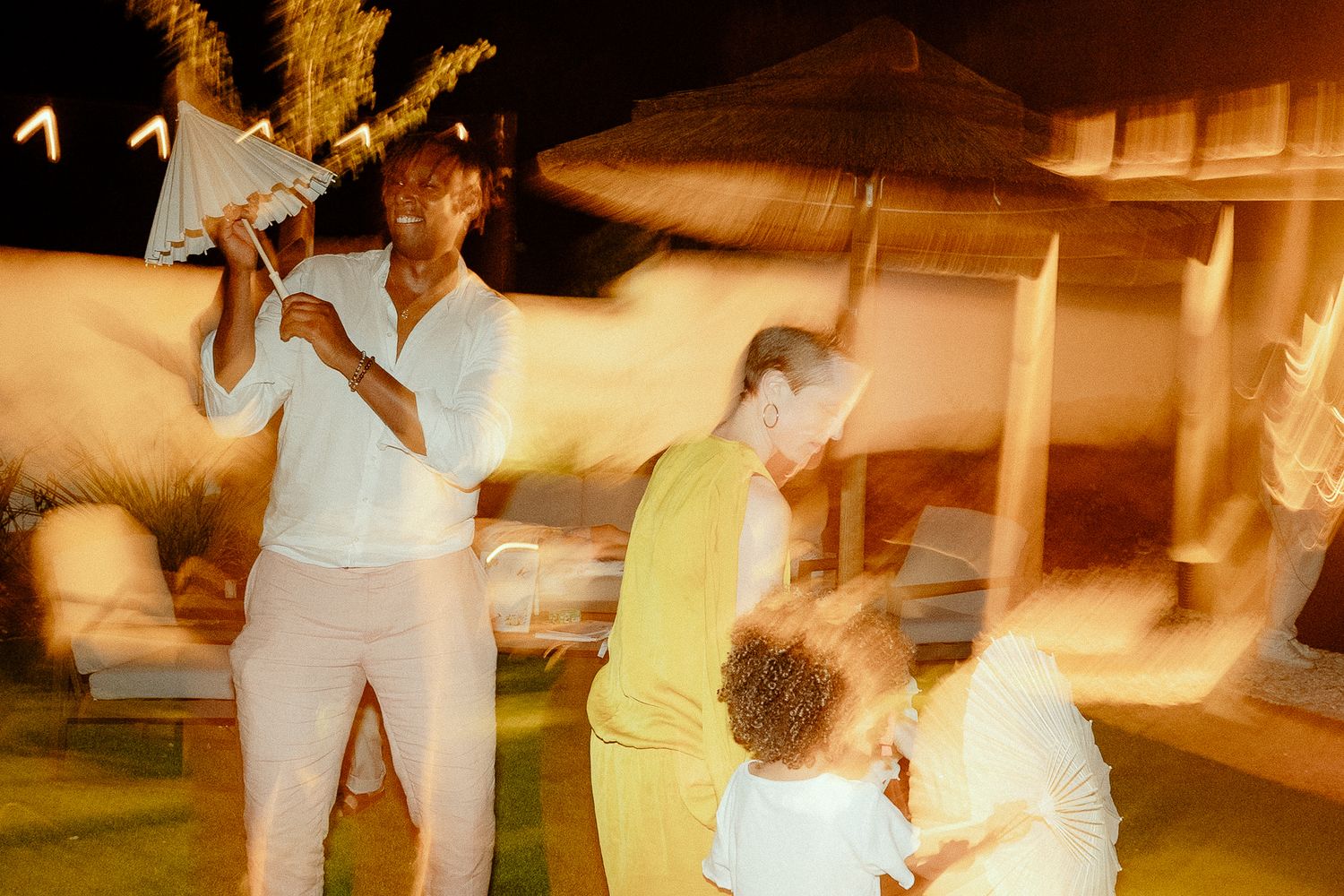 People enjoying a lively outdoor night party with motion blur effects under palm trees and patio umbrellas.