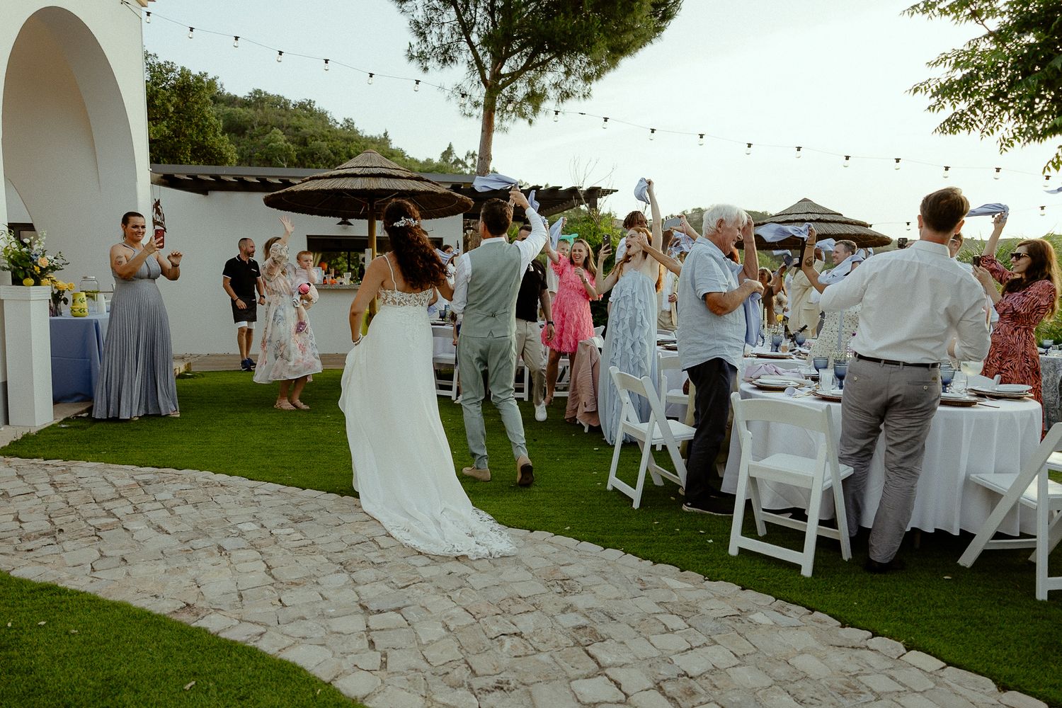 Bride and groom walk through cheering wedding guests at an outdoor garden reception with string lights.