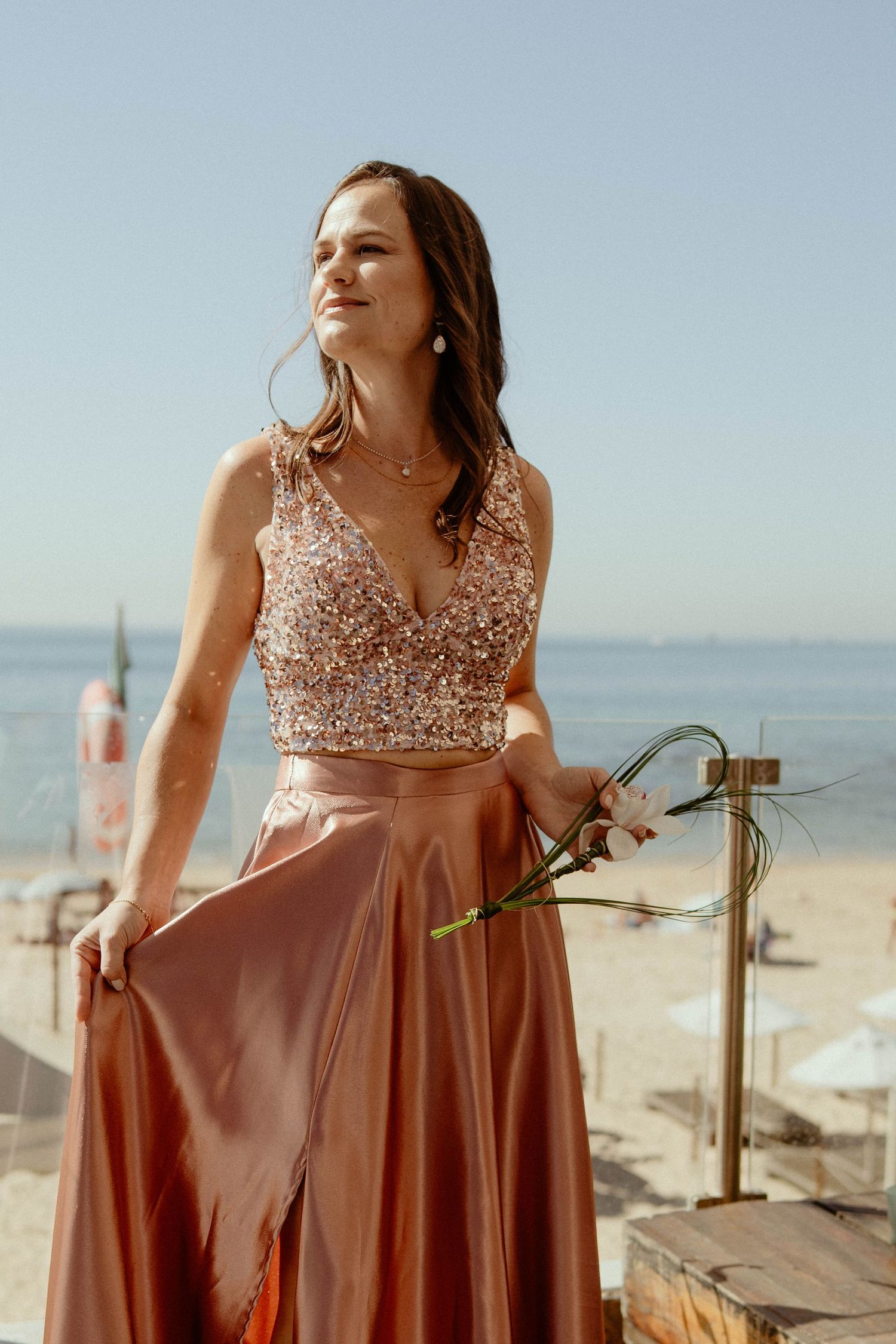 A person in a rose gold sequined dress poses on a beach with the ocean visible in the background.