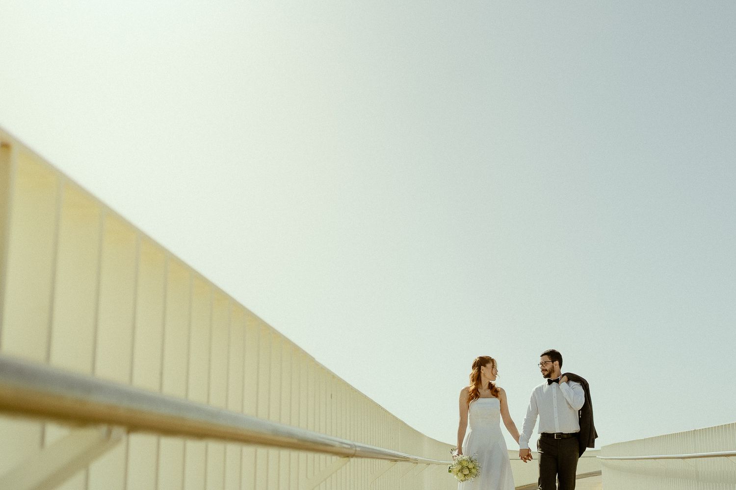Bride and groom holding hands walking on a modern outdoor terrace under a bright clear sky on their wedding day.