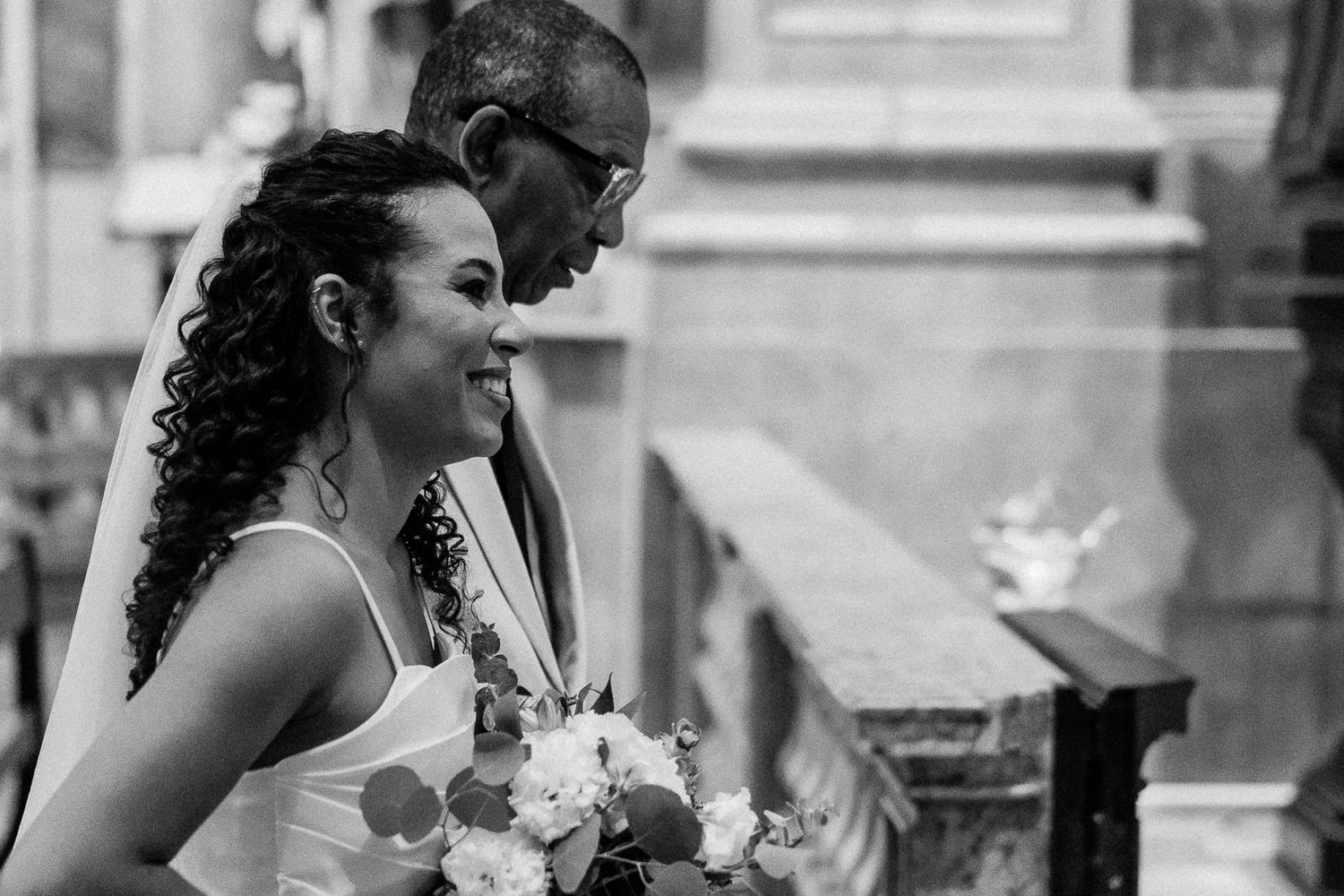 Black and white wedding photo of a smiling bride holding bouquet with groom leaning in close inside a church.