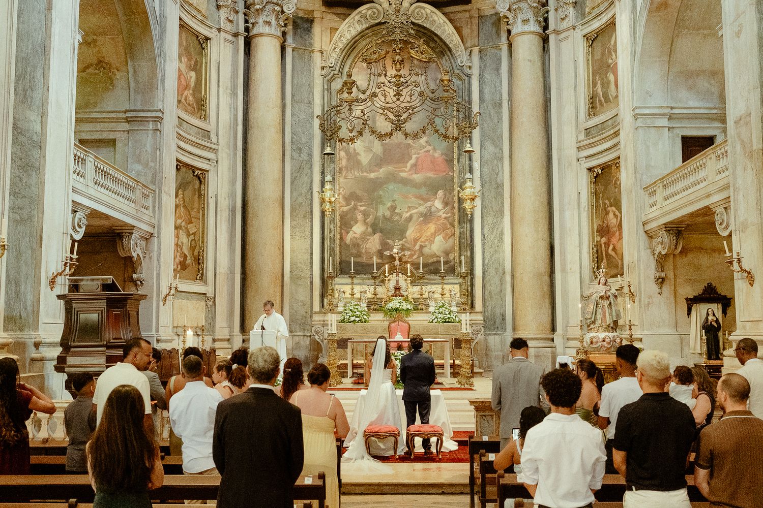 Wedding ceremony at ornate baroque church altar with guests seated in pews, bride and groom standing before priest.