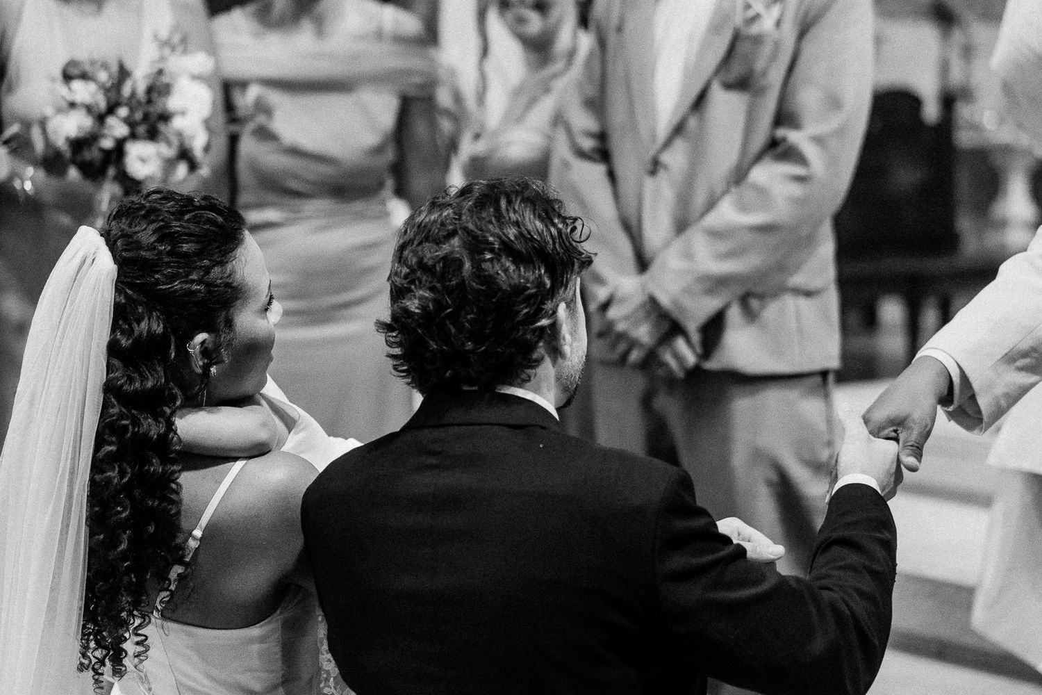 Black and white wedding ceremony photo showing bride and groom kneeling at altar with guests standing behind them.