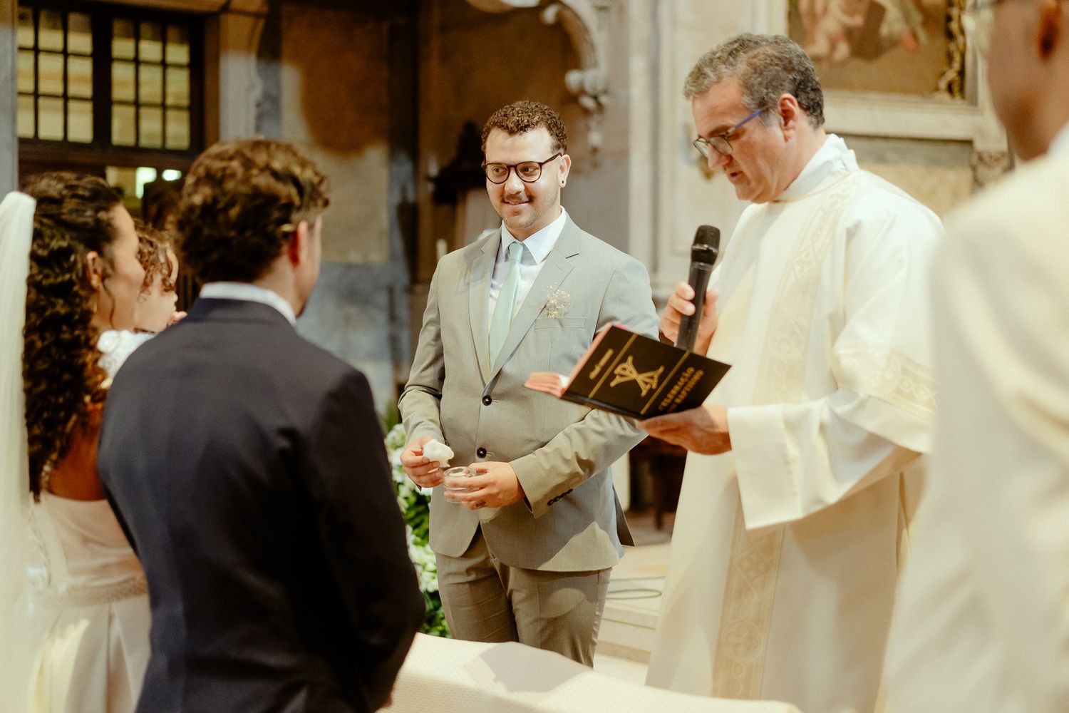 Groom in gray suit smiling during wedding ceremony with officiant holding microphone and book.