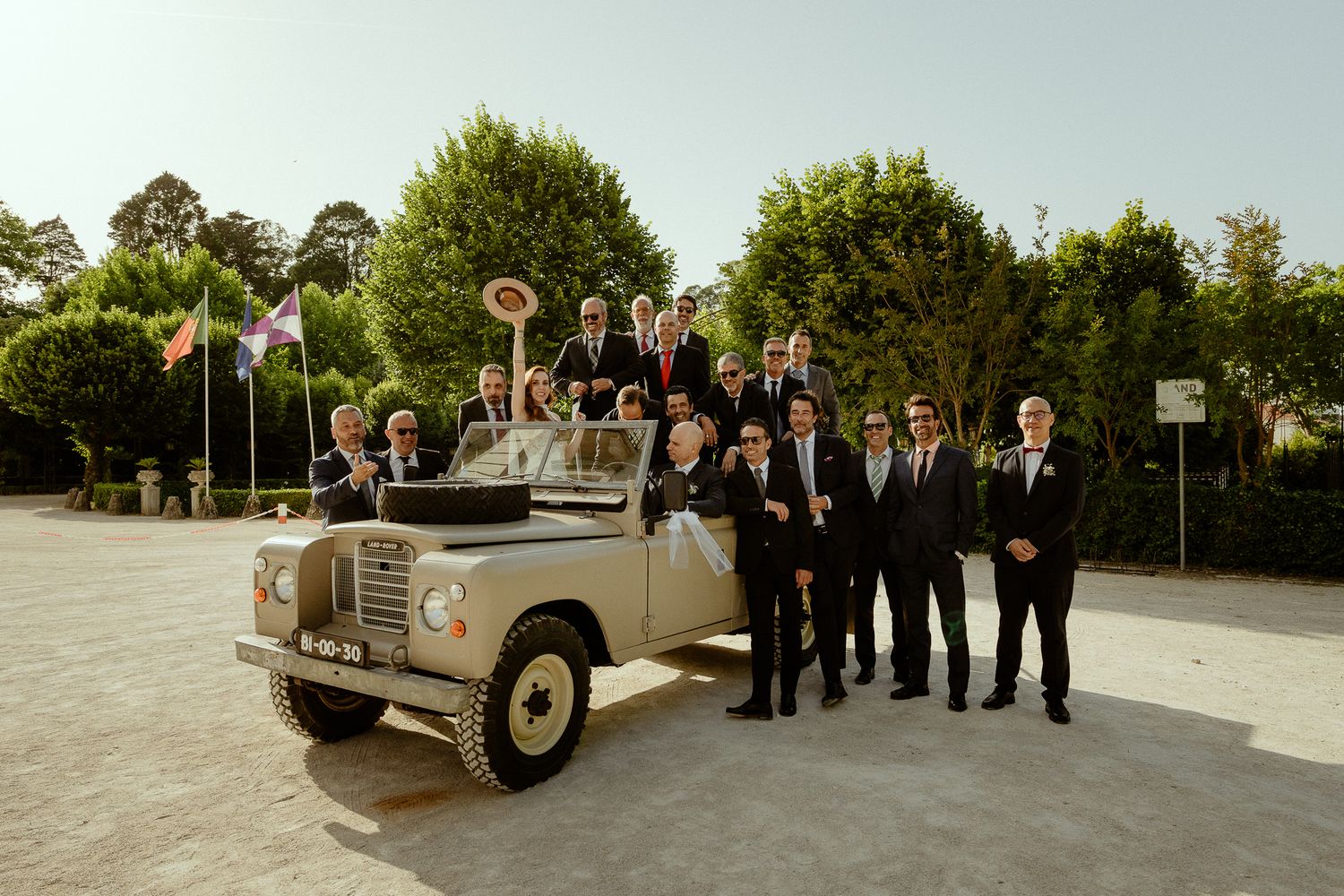Group of men in suits posing beside a vintage Land Rover at an outdoor venue with trees and flags.