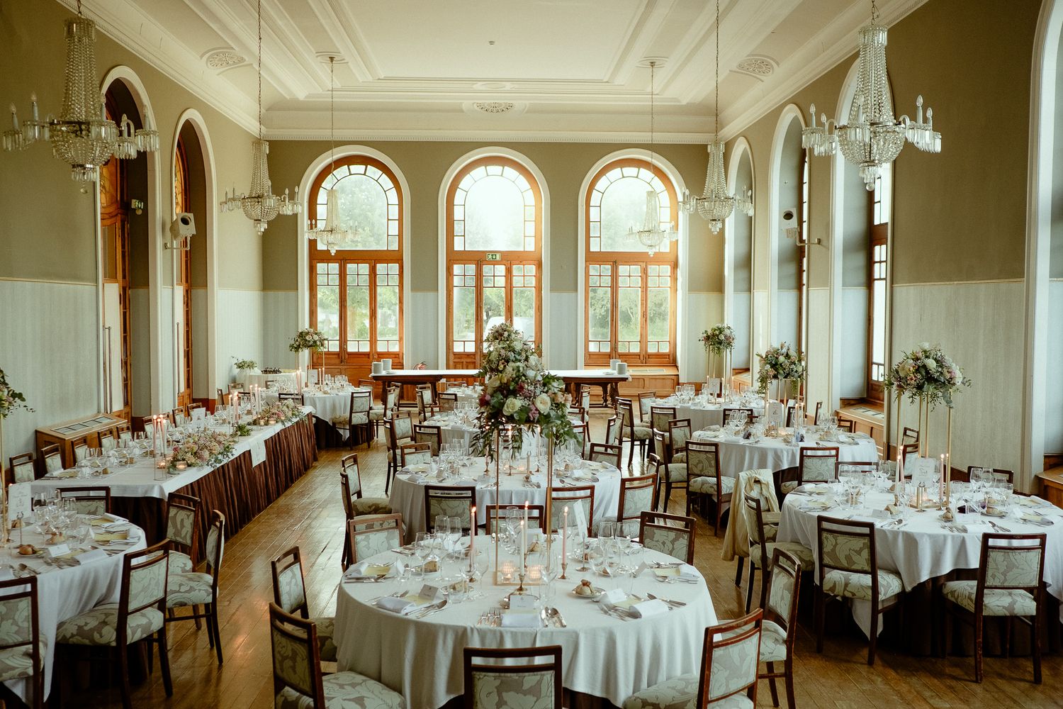 Elegant wedding reception hall with round tables, white linens, floral centerpieces, chandeliers, and arched windows.