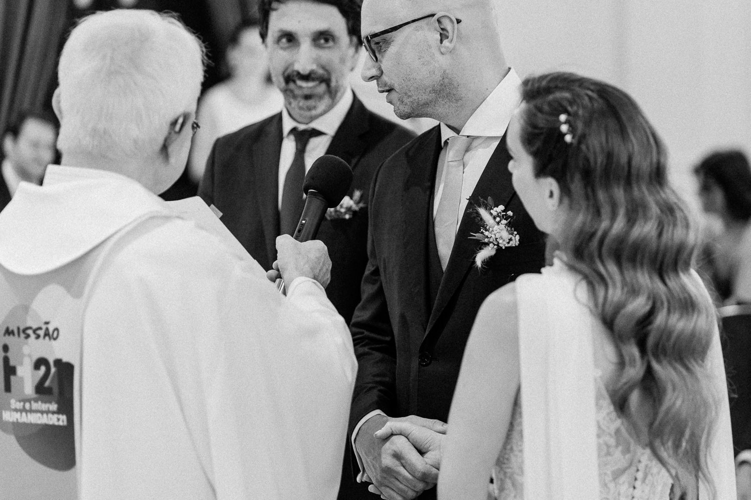 Black and white wedding ceremony photo with bride, groom, best man, and officiant exchanging vows in a church.