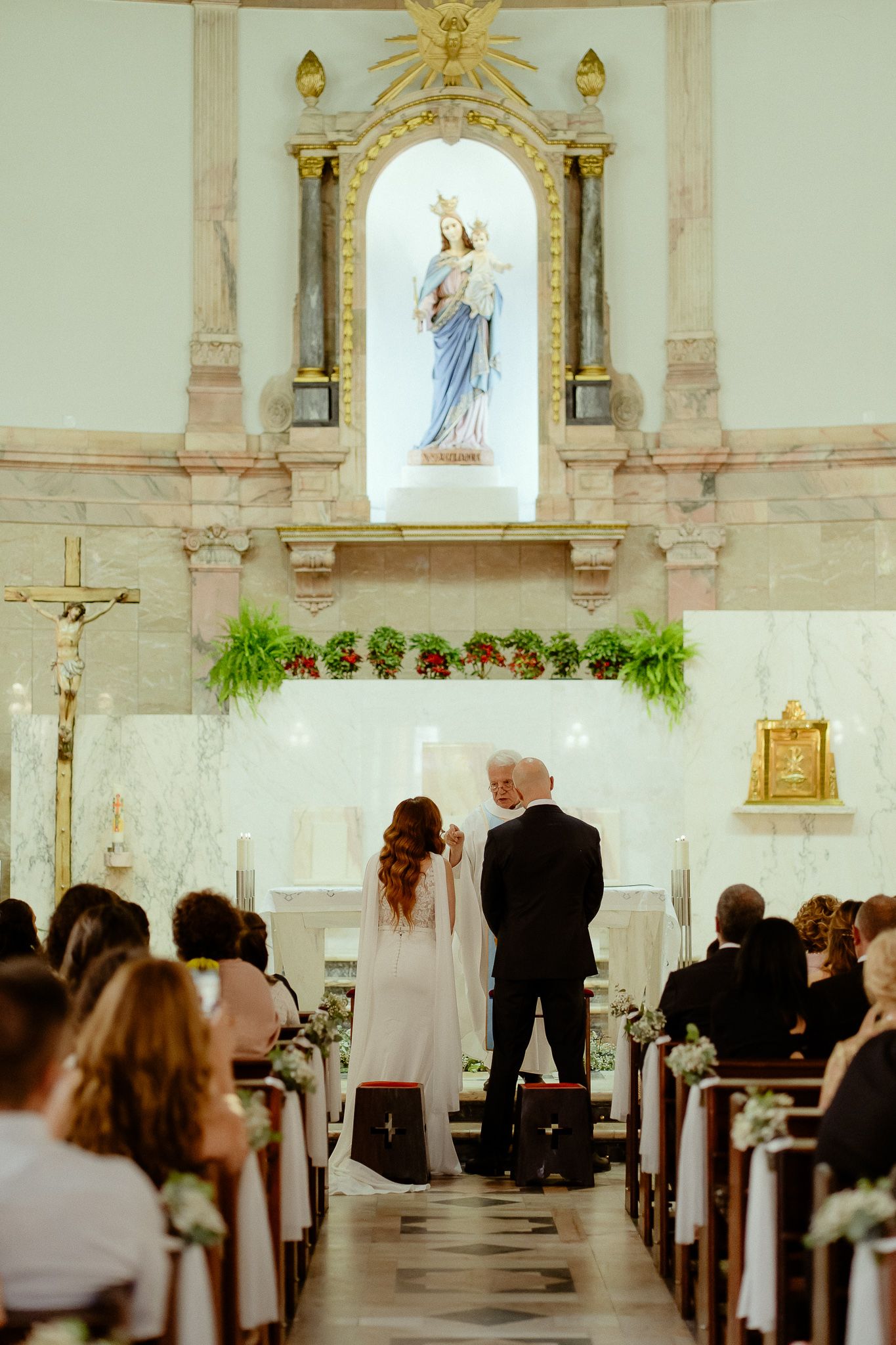 Bride and groom stand at church altar during wedding ceremony with Virgin Mary statue and floral arrangements.