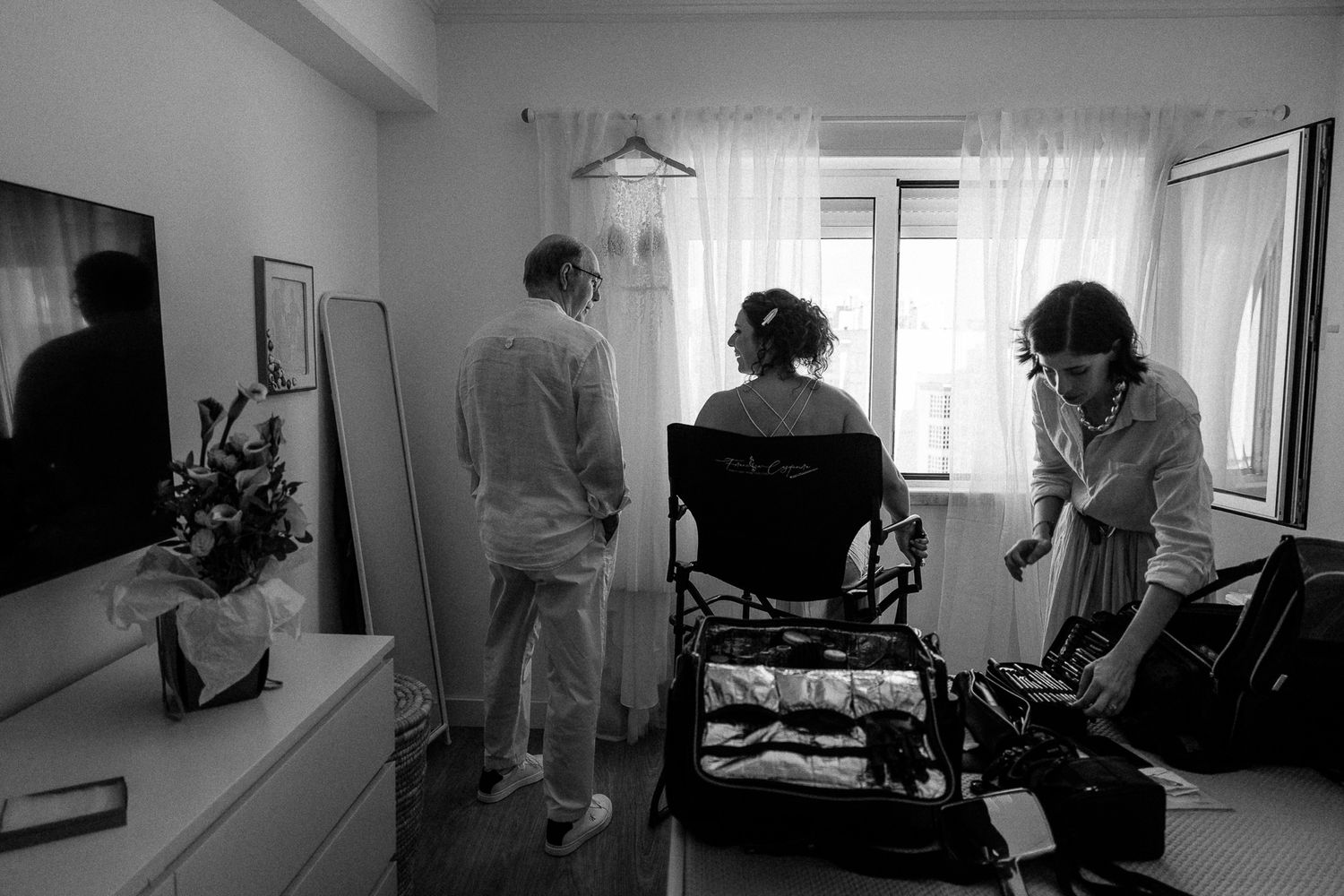 Black and white photo of three people in a hotel room, one seated in a chair, preparing for a wedding ceremony.