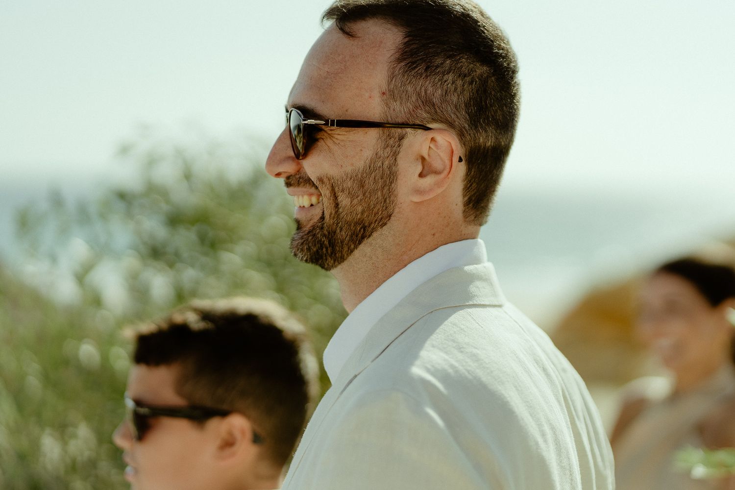 Smiling man with beard and sunglasses in white shirt at outdoor wedding ceremony on sunny day.