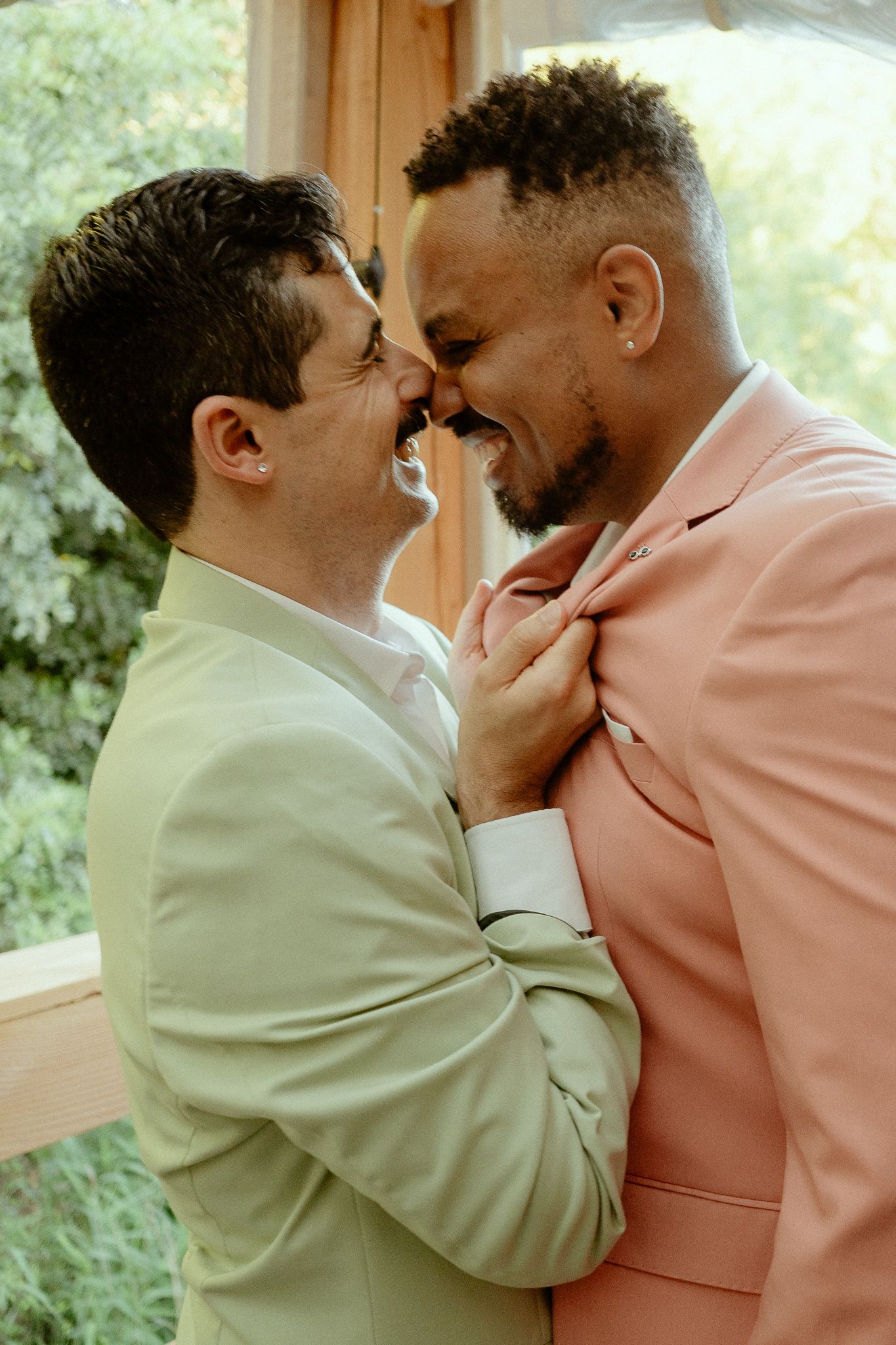 Two men in pastel suits share a tender moment, smiling and embracing near a window with greenery.