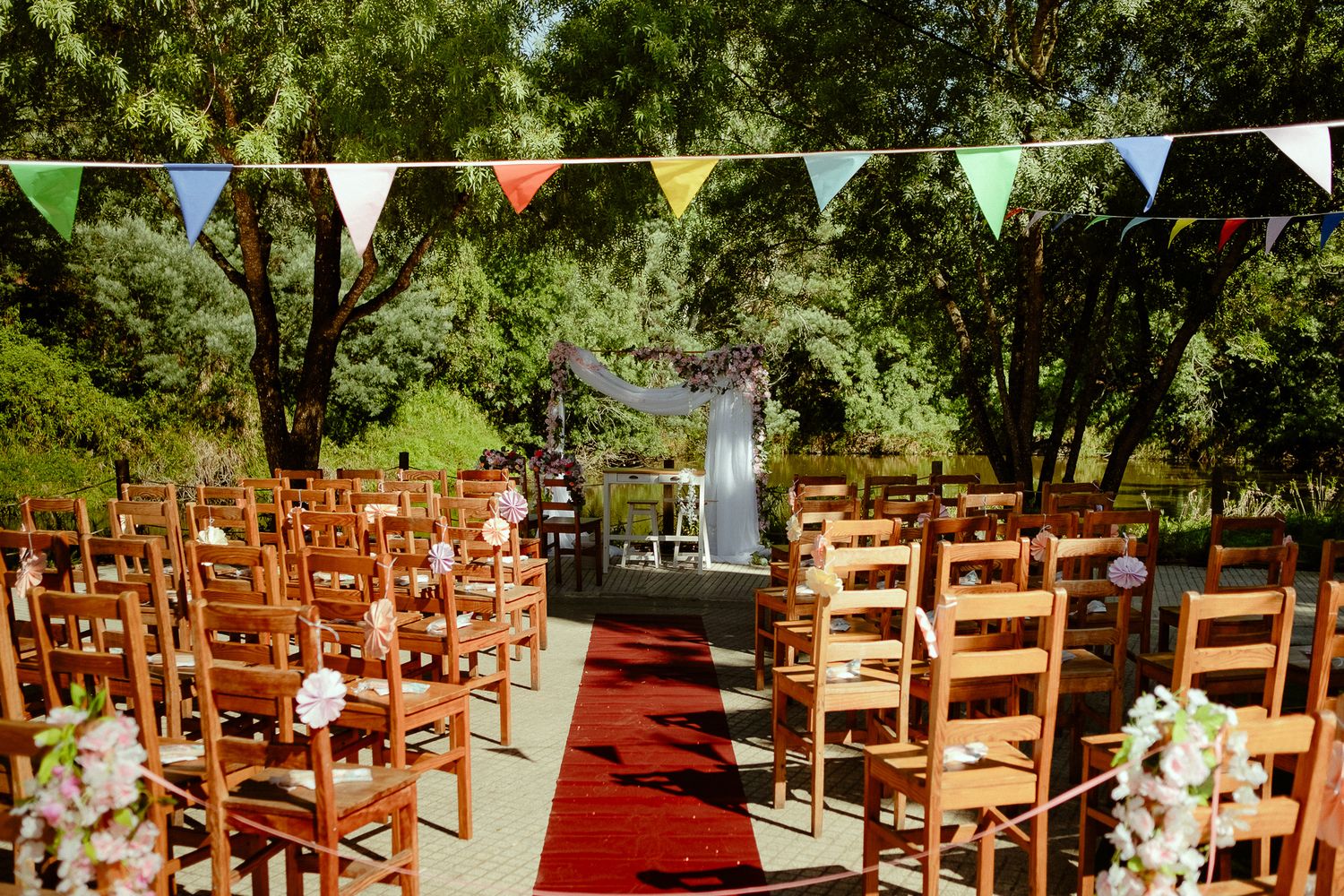 Outdoor wedding ceremony setup with wooden chairs, red carpet aisle, colorful bunting flags, and floral arch under shady trees.