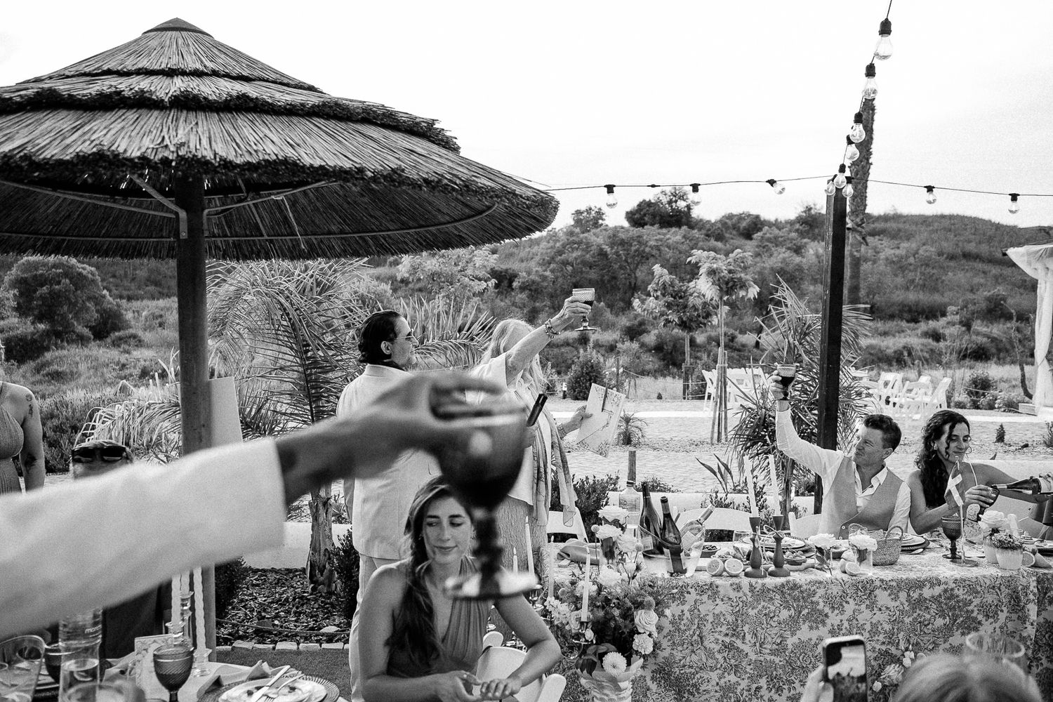 Black and white outdoor wedding reception toast with guests gathered under thatched umbrellas and string lights.
