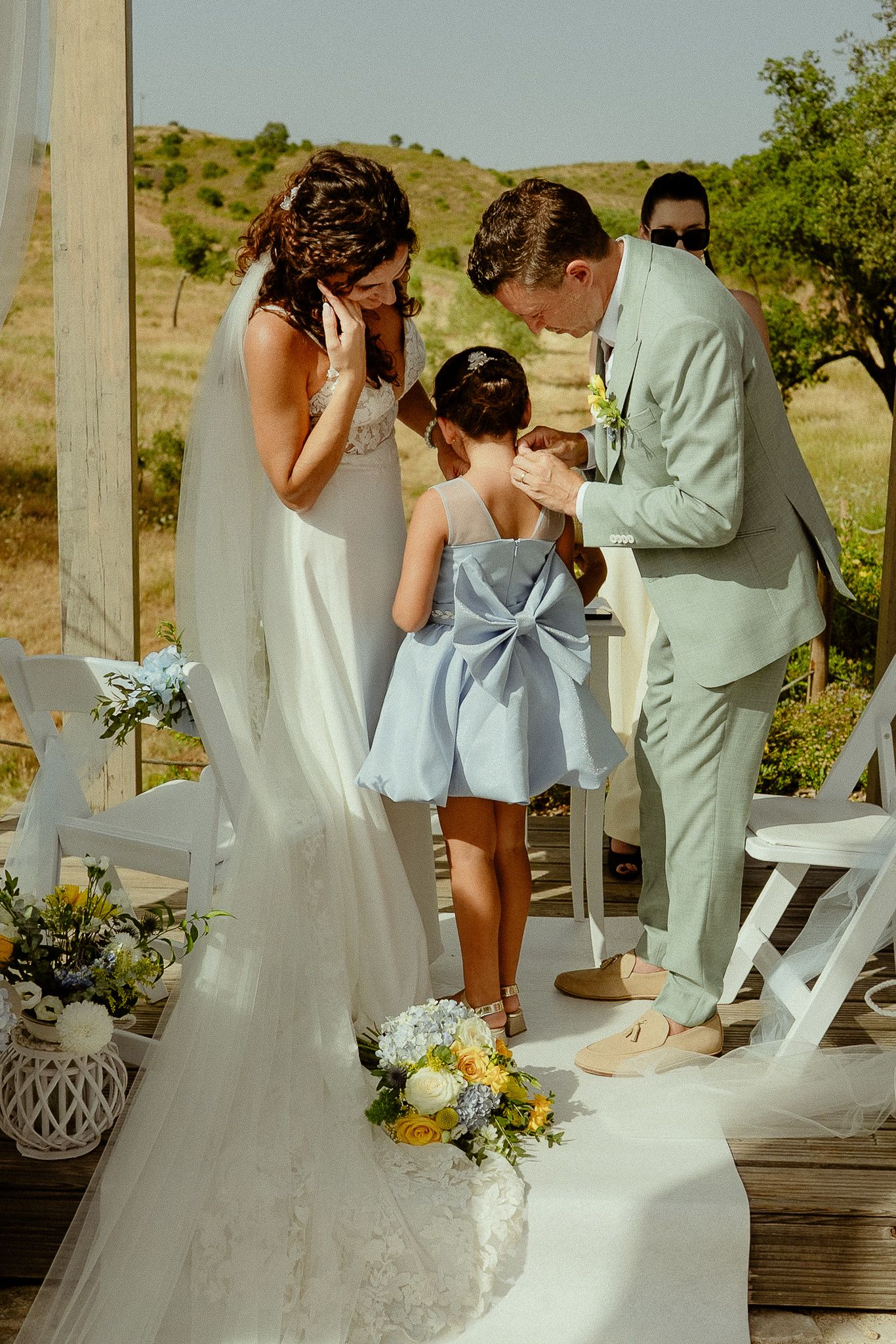 Bride and groom share a tender moment with flower girl in blue bow dress at outdoor wedding ceremony.