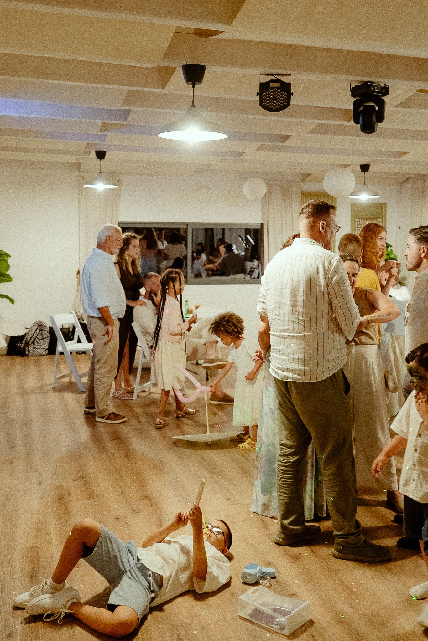 Group of adults and children gathered in a bright indoor venue with wooden floors during a casual social event.