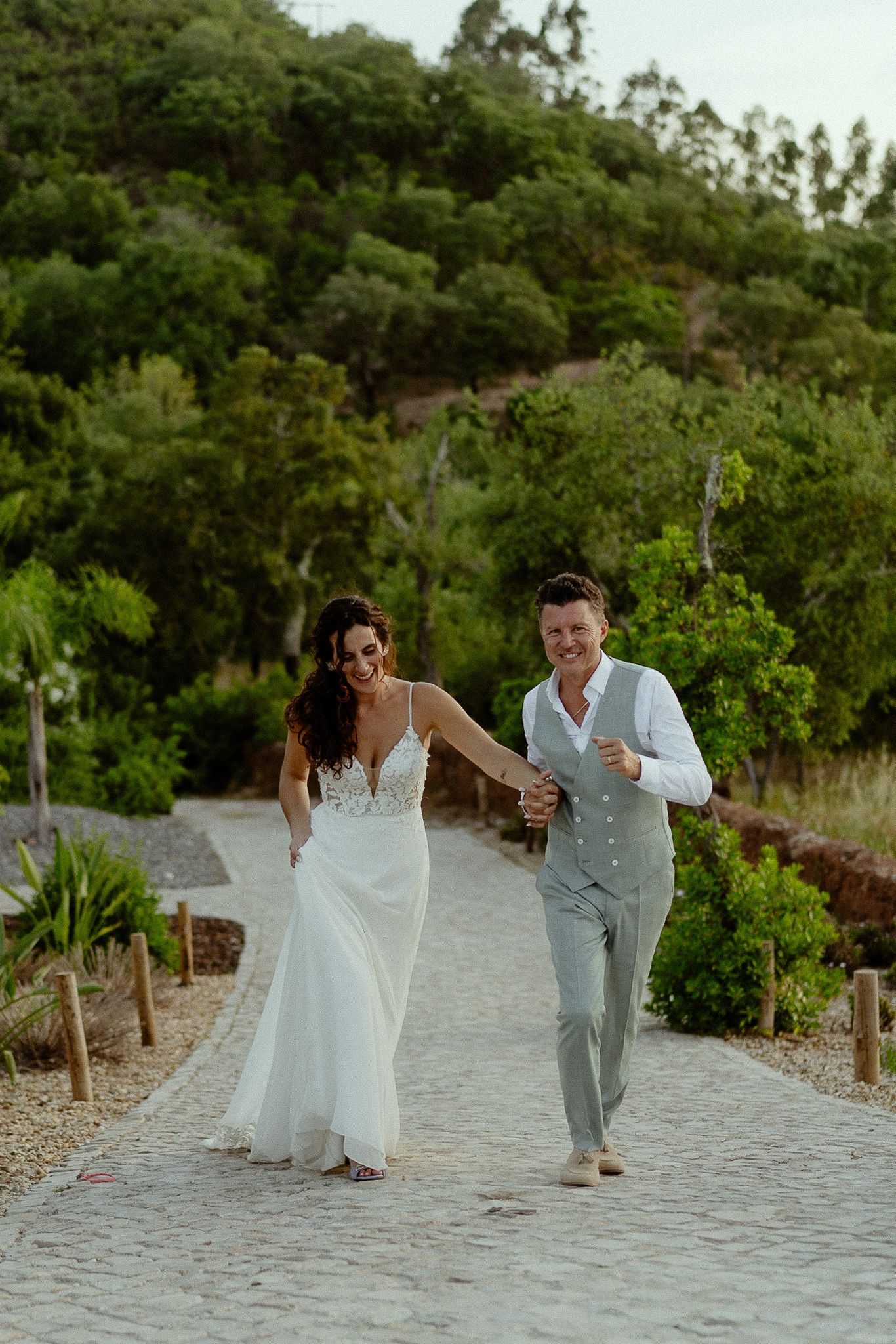 A joyful bride and groom walk hand-in-hand along a stone path surrounded by lush greenery on their wedding day.