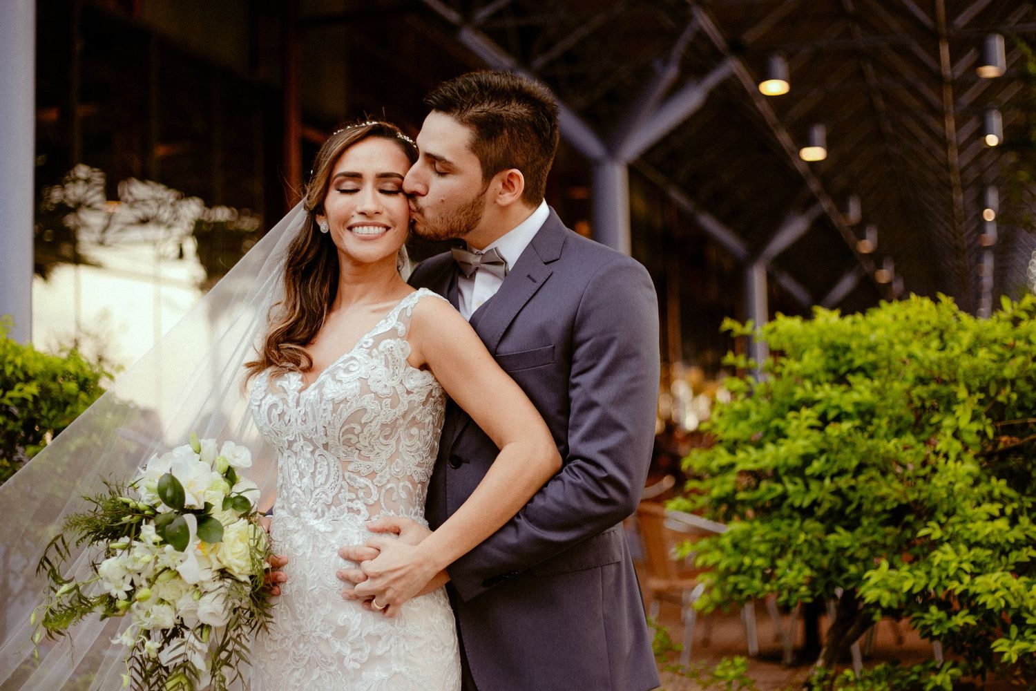 A couple in wedding attire share a romantic moment outdoors near flowering plants and greenery.