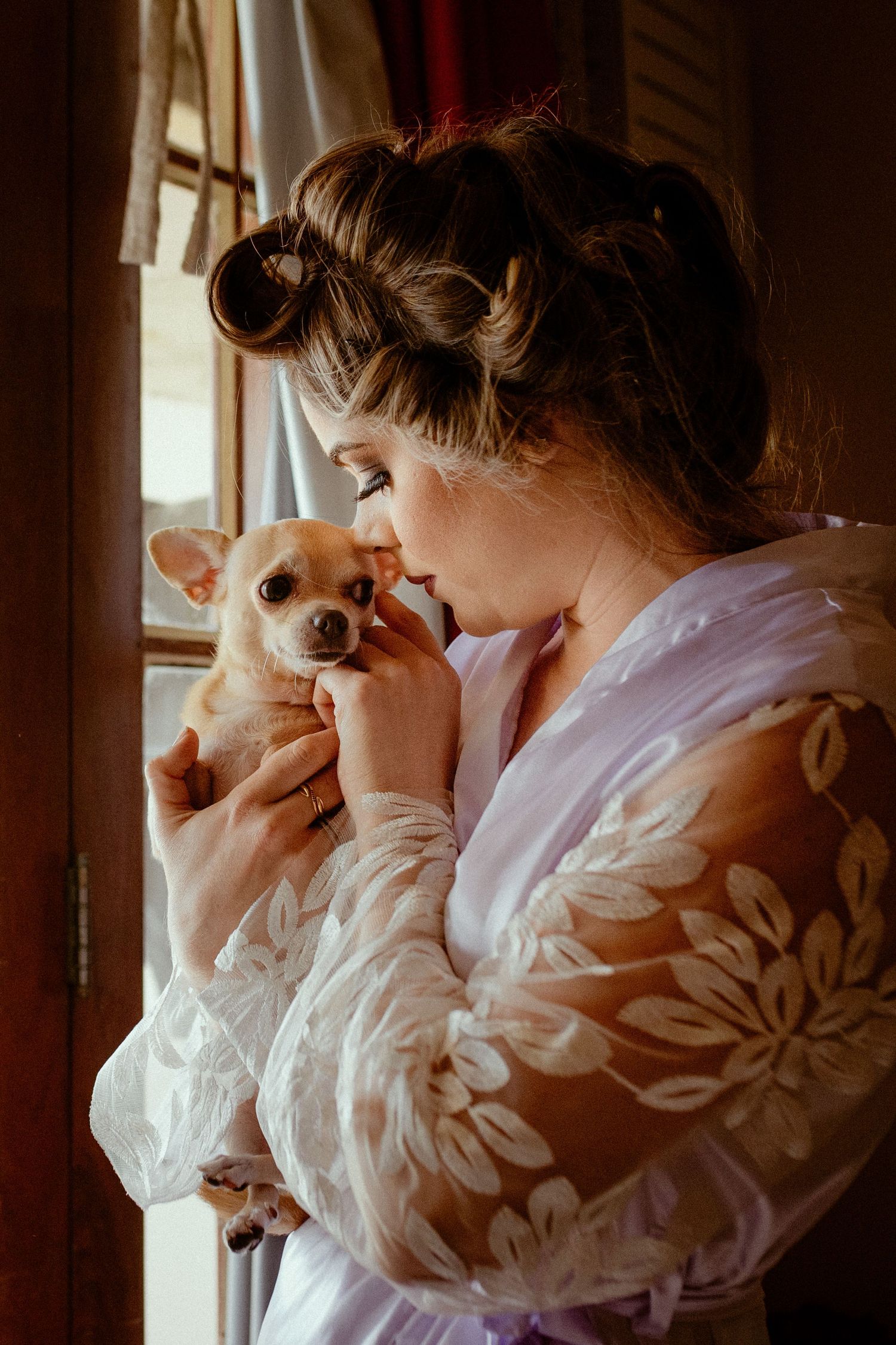 A person in a white floral robe looks through window blinds in warm lighting.