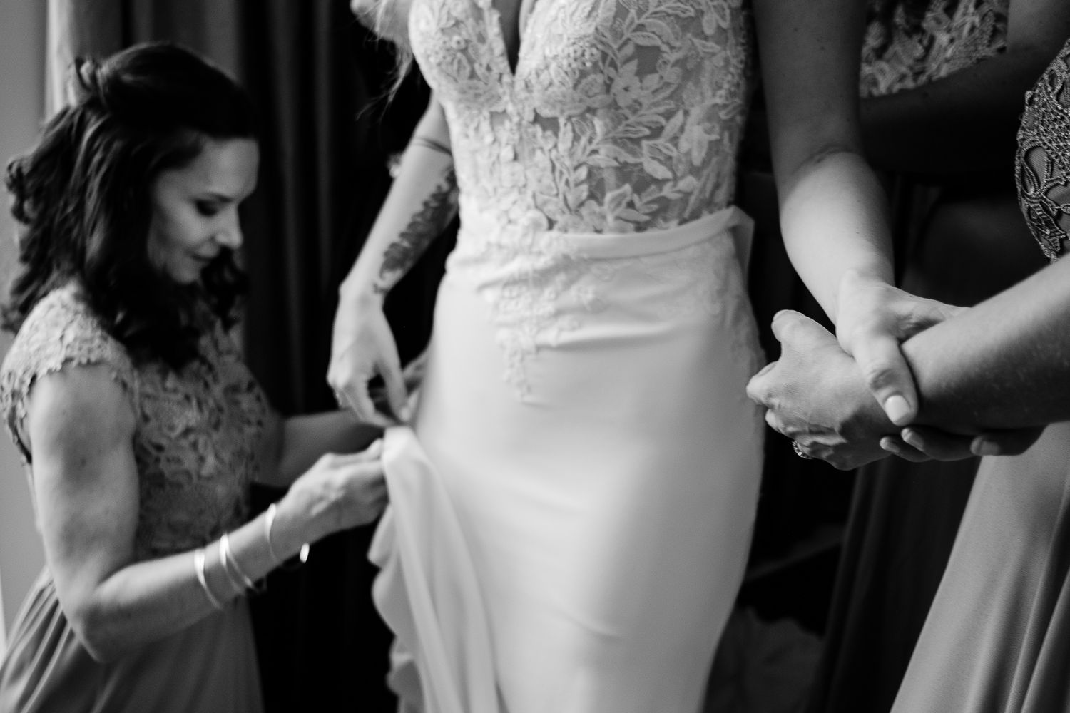 Bridesmaids helping bride into elegant lace wedding gown in black and white photo.