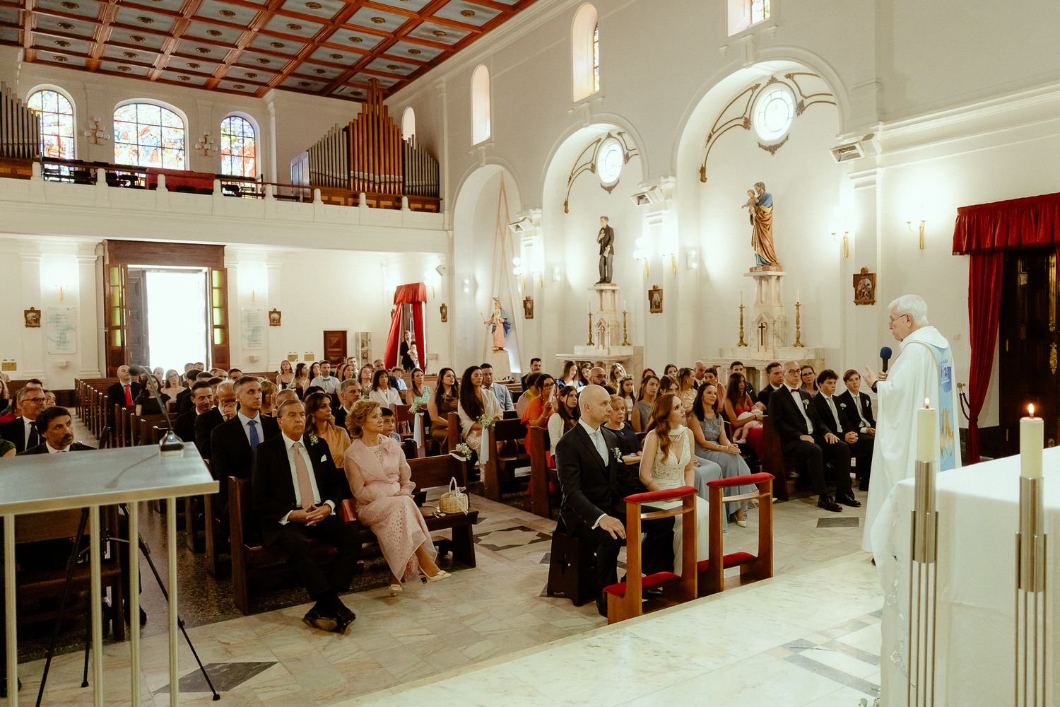 Guests gathered in an elegant white church for a wedding ceremony with a priest officiating at the altar.