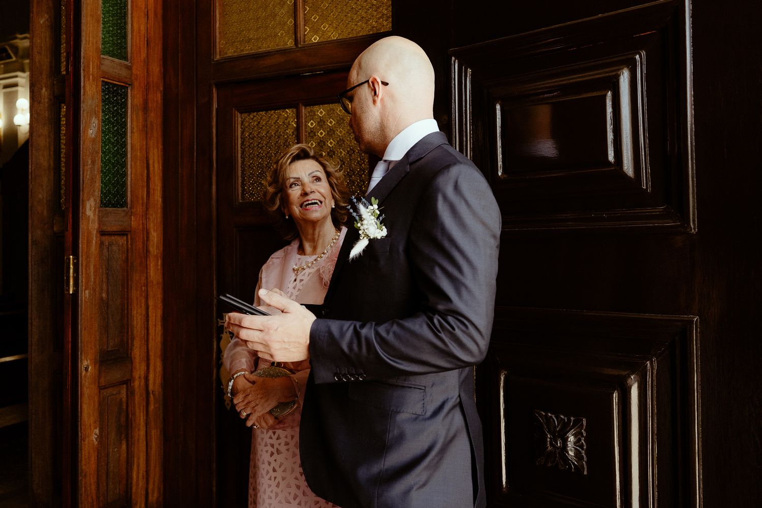 Groom in navy suit with boutonniere speaks with elegant older woman in pink dress near ornate wooden doors.