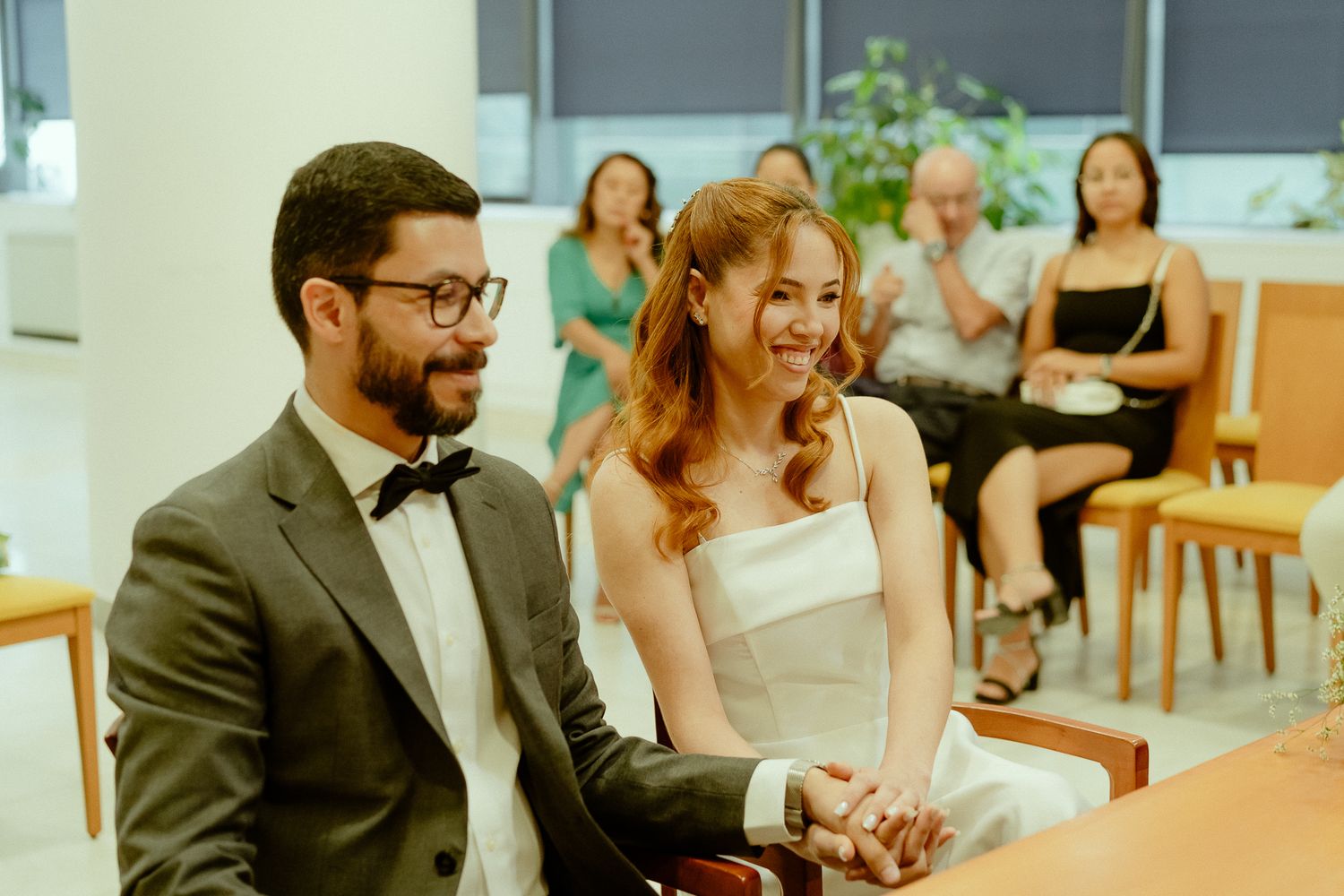 Happy couple holding hands at their wedding ceremony, groom in gray suit, bride in white dress, guests seated behind.