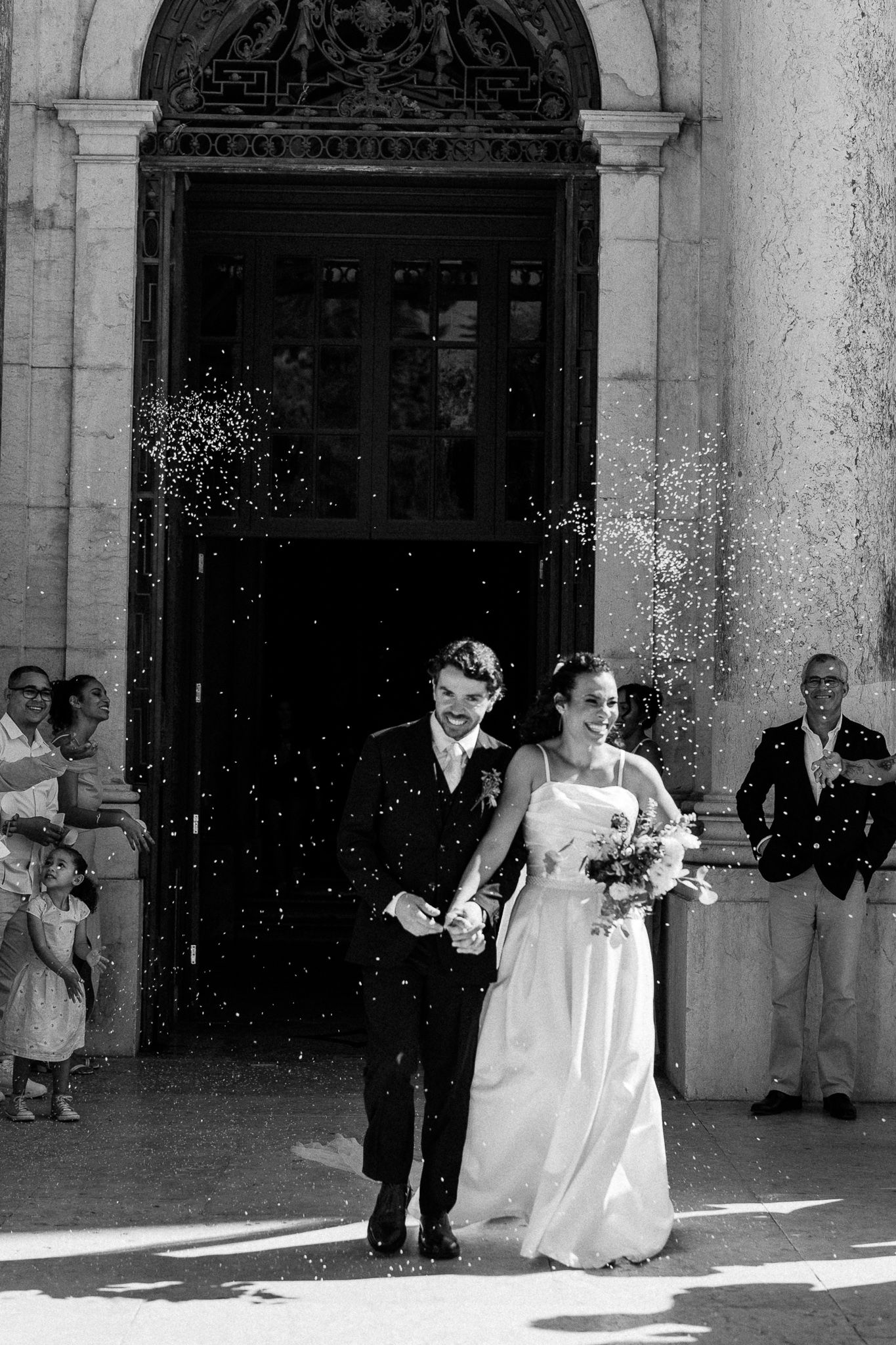 Black and white photo of a newlywed couple exiting a church amid a shower of rice, surrounded by guests.