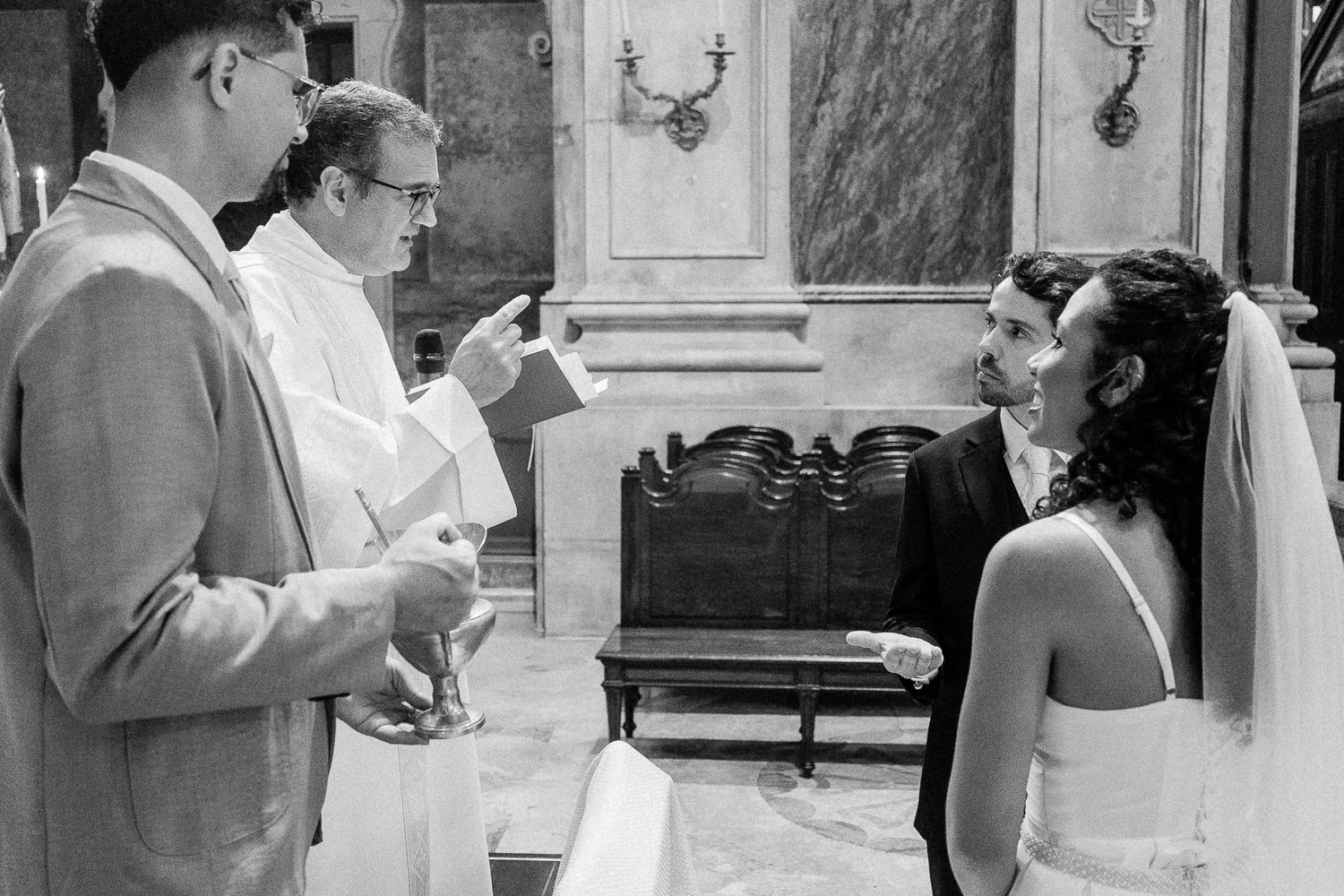 Black and white wedding ceremony photo with bride, groom, and officiant exchanging vows in elegant church.