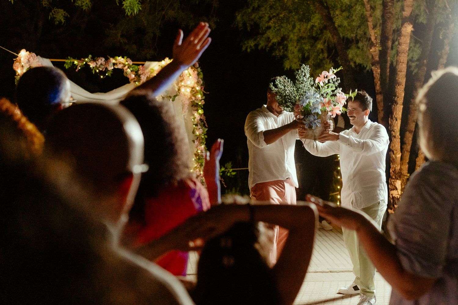 Two people hold a floral bouquet together at an outdoor nighttime wedding celebration with guests cheering.