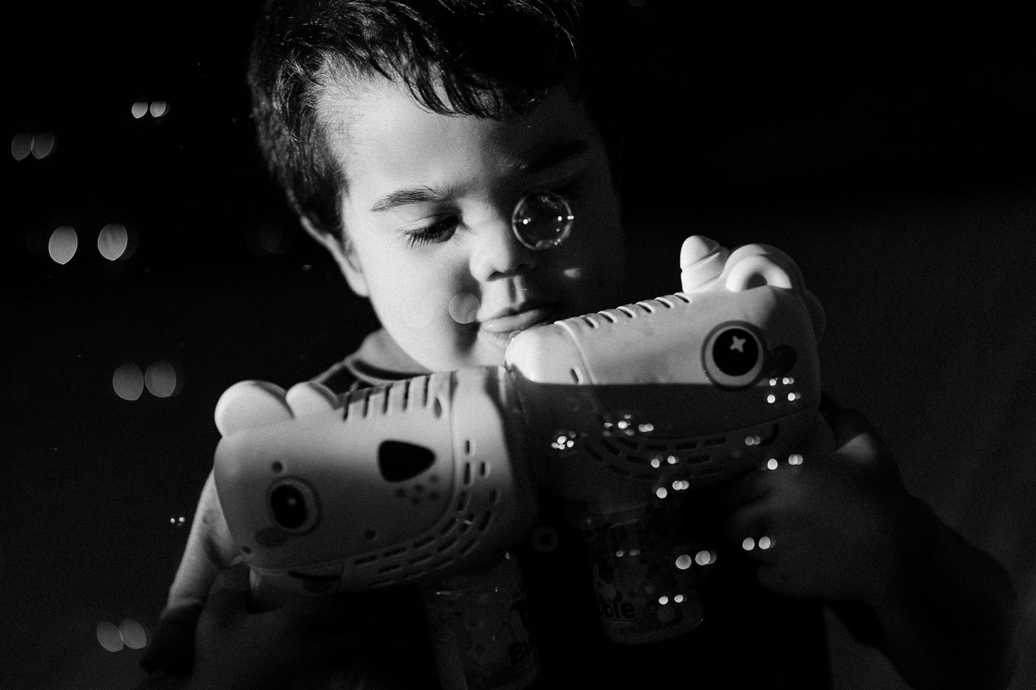 Black and white photo of a young boy holding and examining two stuffed animal toys in dim lighting.