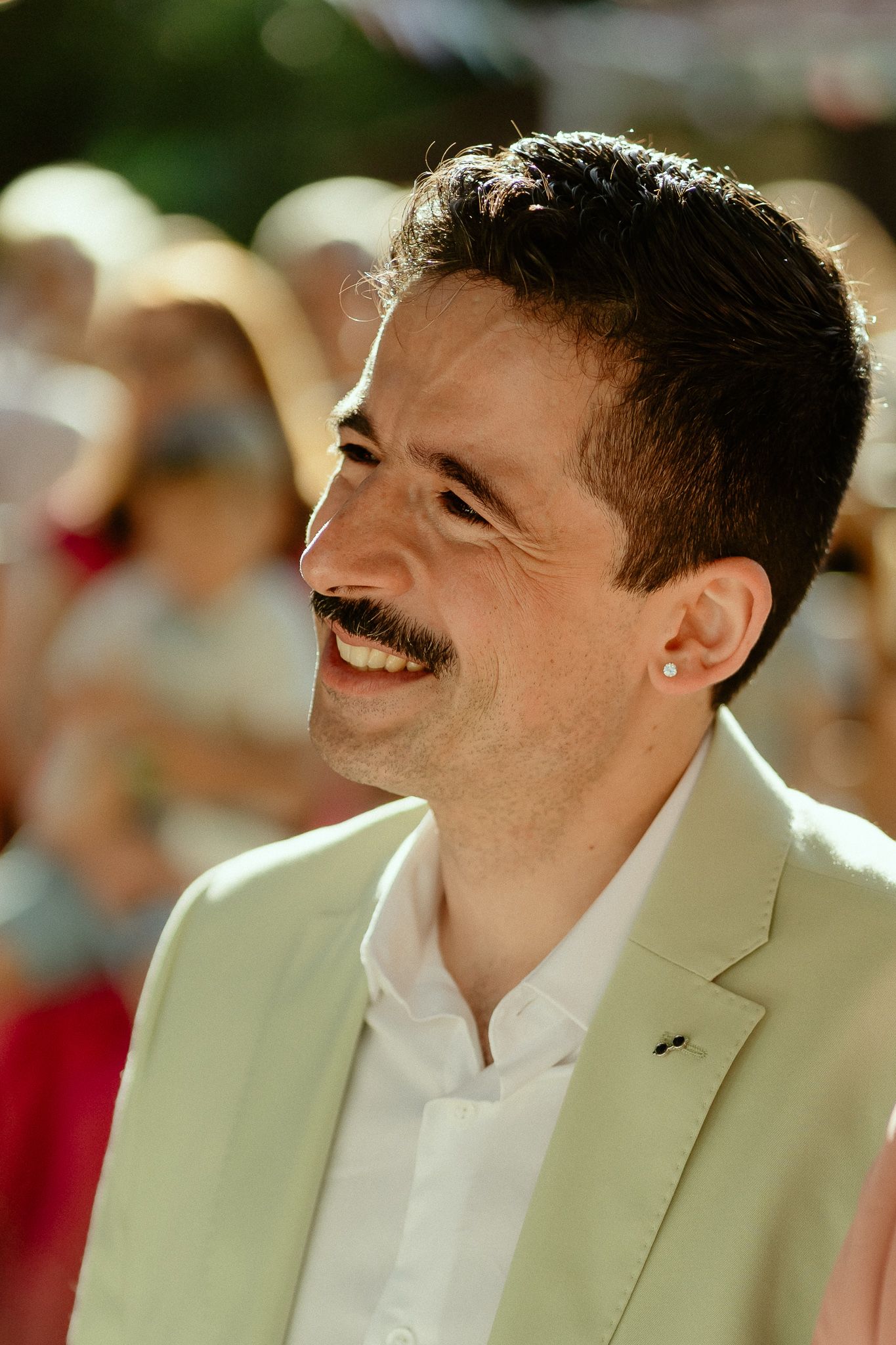 Smiling man with mustache wearing white blazer and earring at outdoor event in sunlight.
