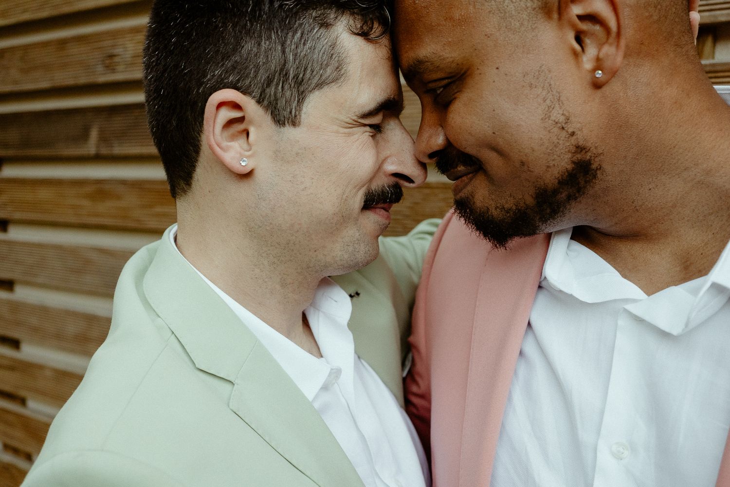 Two men in pastel suits share an intimate forehead-to-forehead moment, smiling warmly at each other.