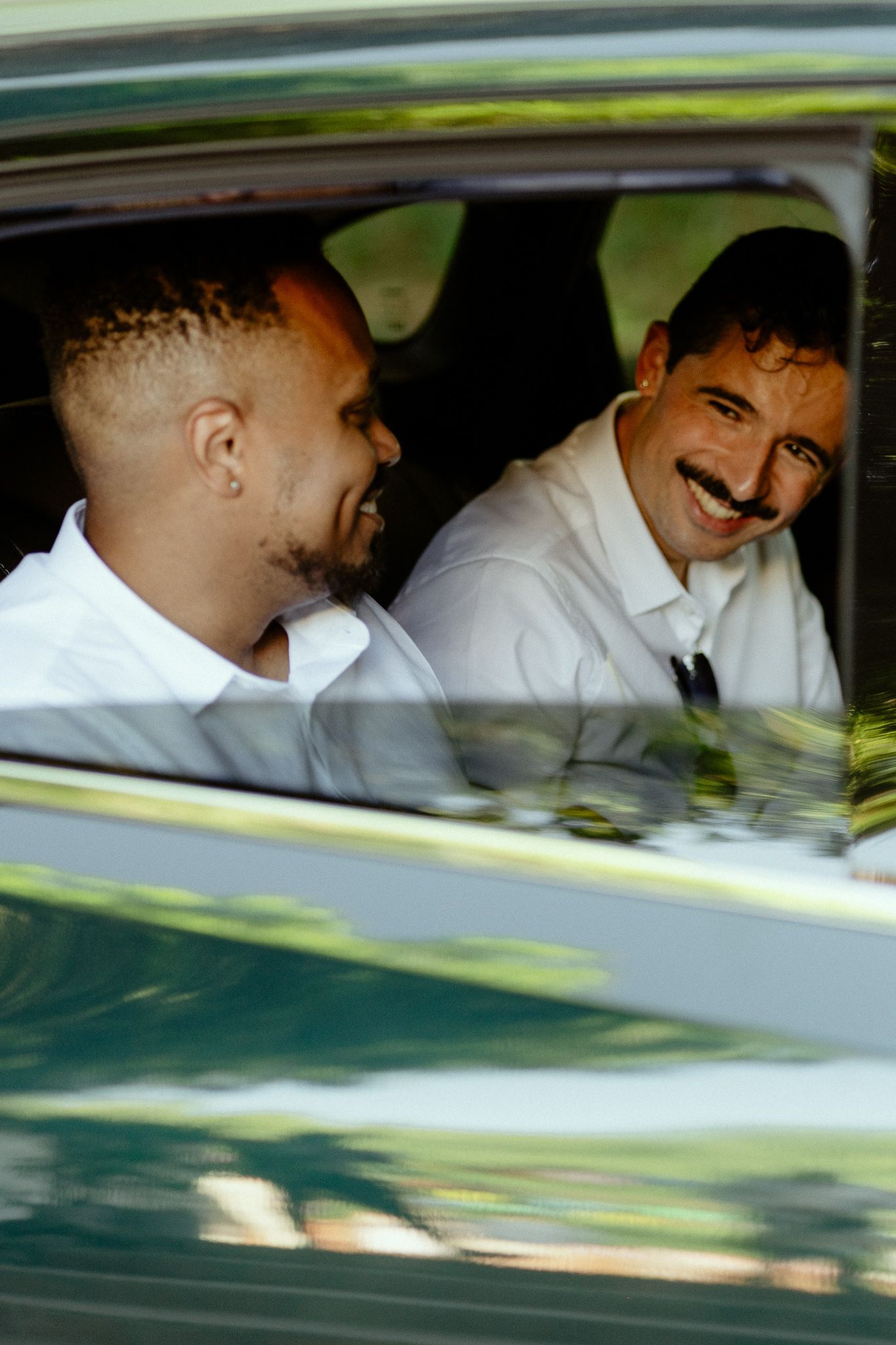 Two men in white shirts smiling and laughing together inside a vintage car.