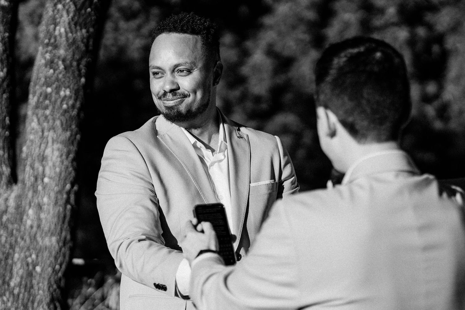 Black and white photo of two men in suits sharing a handshake outdoors at a wedding ceremony.