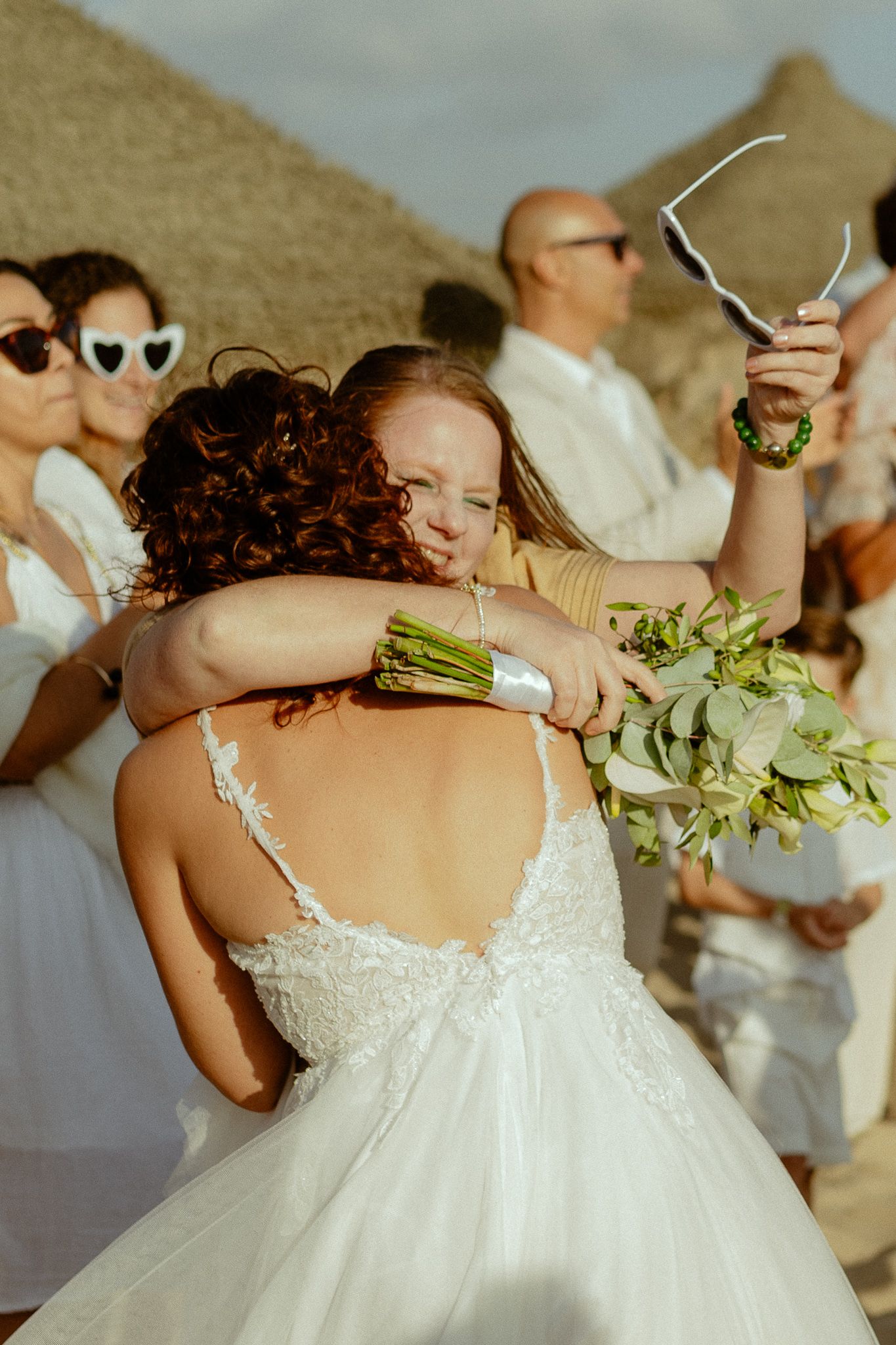 Bride in backless lace wedding dress embracing guest while holding floral bouquet at outdoor beach wedding ceremony.