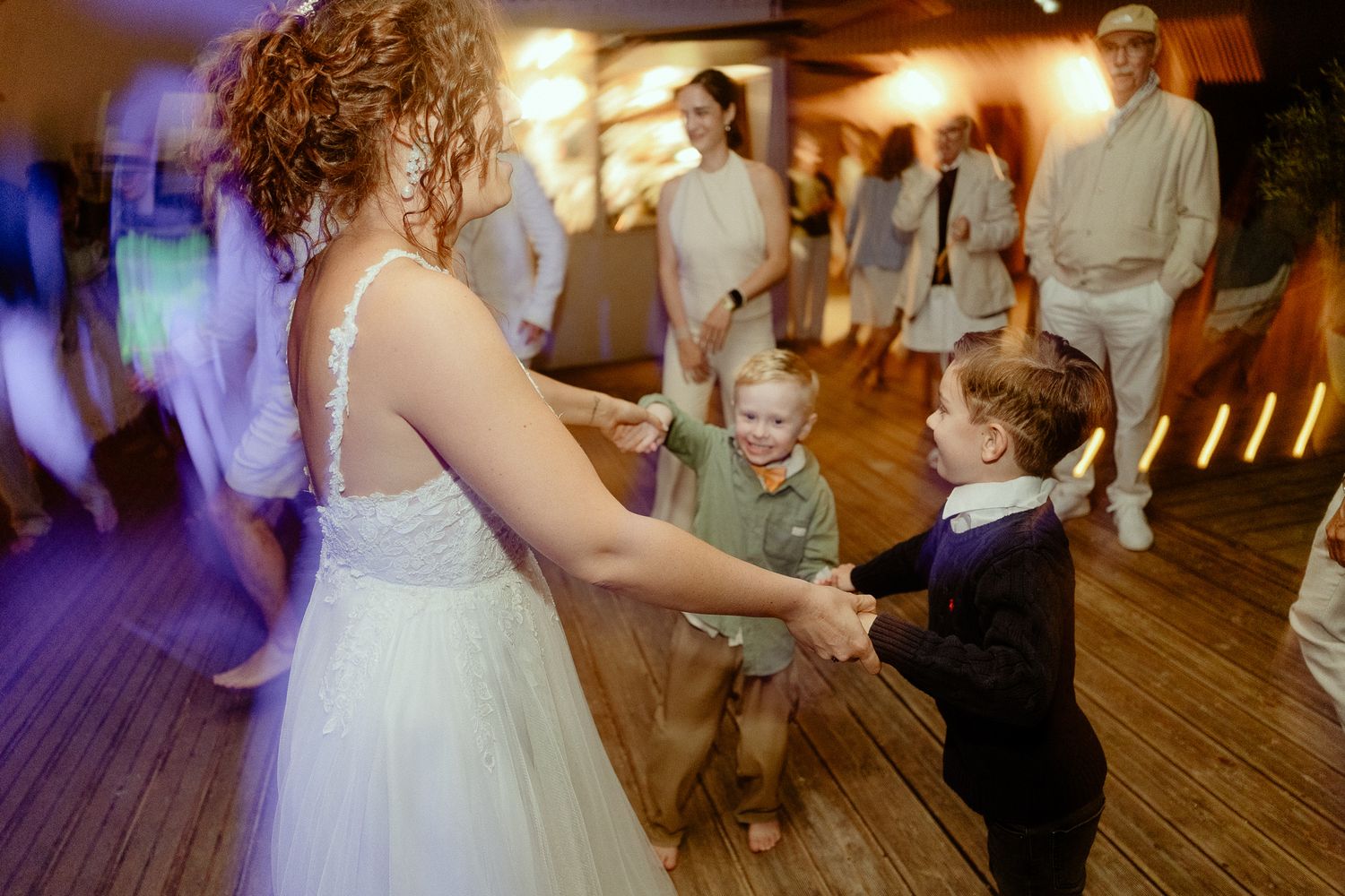 Bride in white gown dancing with young children on wedding reception dance floor with colorful lights.