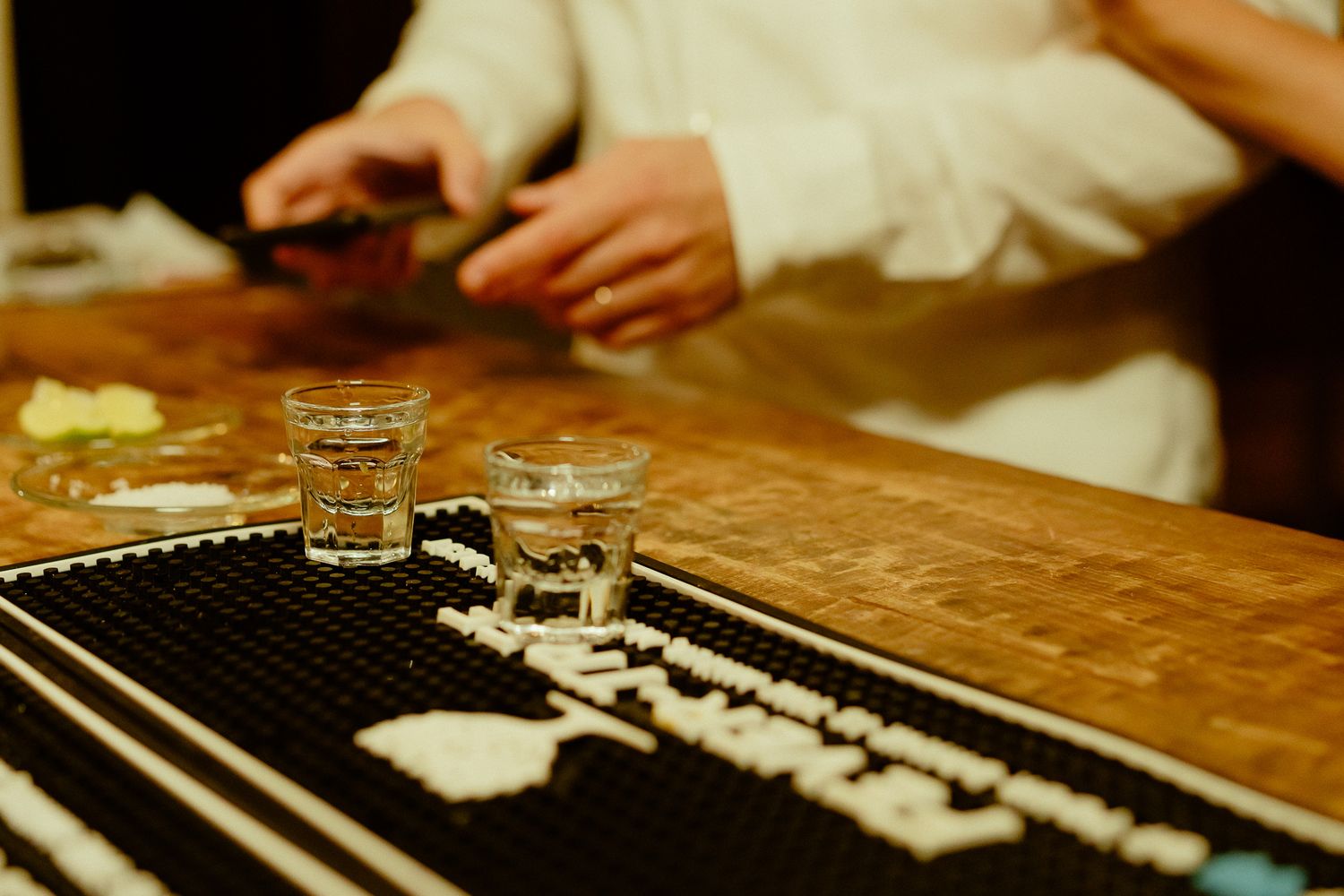 Shot glasses and chopsticks on a wooden bar counter with a person handling food in the background.