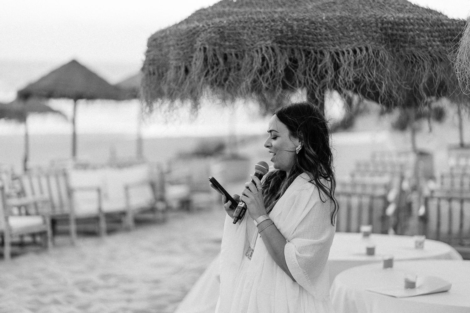 Woman in white dress speaks into microphone at elegant beach wedding reception with thatched umbrellas.