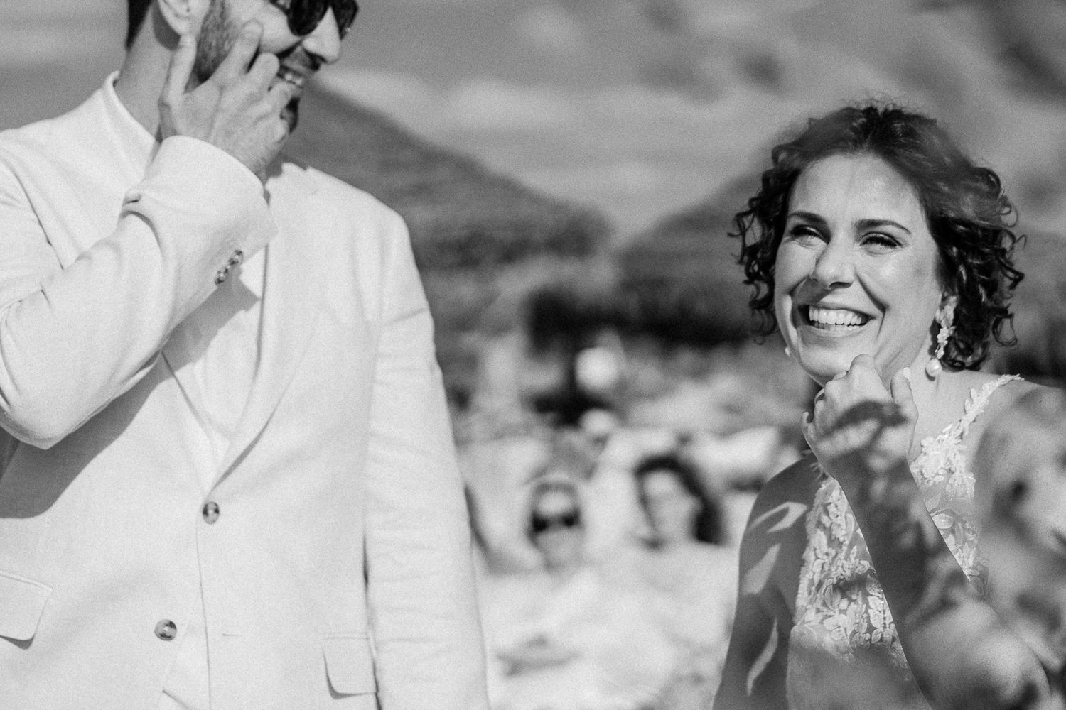 Black and white wedding photo of a laughing bride and groom in white suit during outdoor ceremony.
