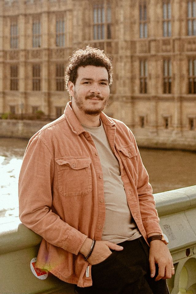 A person in a coral-colored jacket leans against a railing with historic architecture in the background.