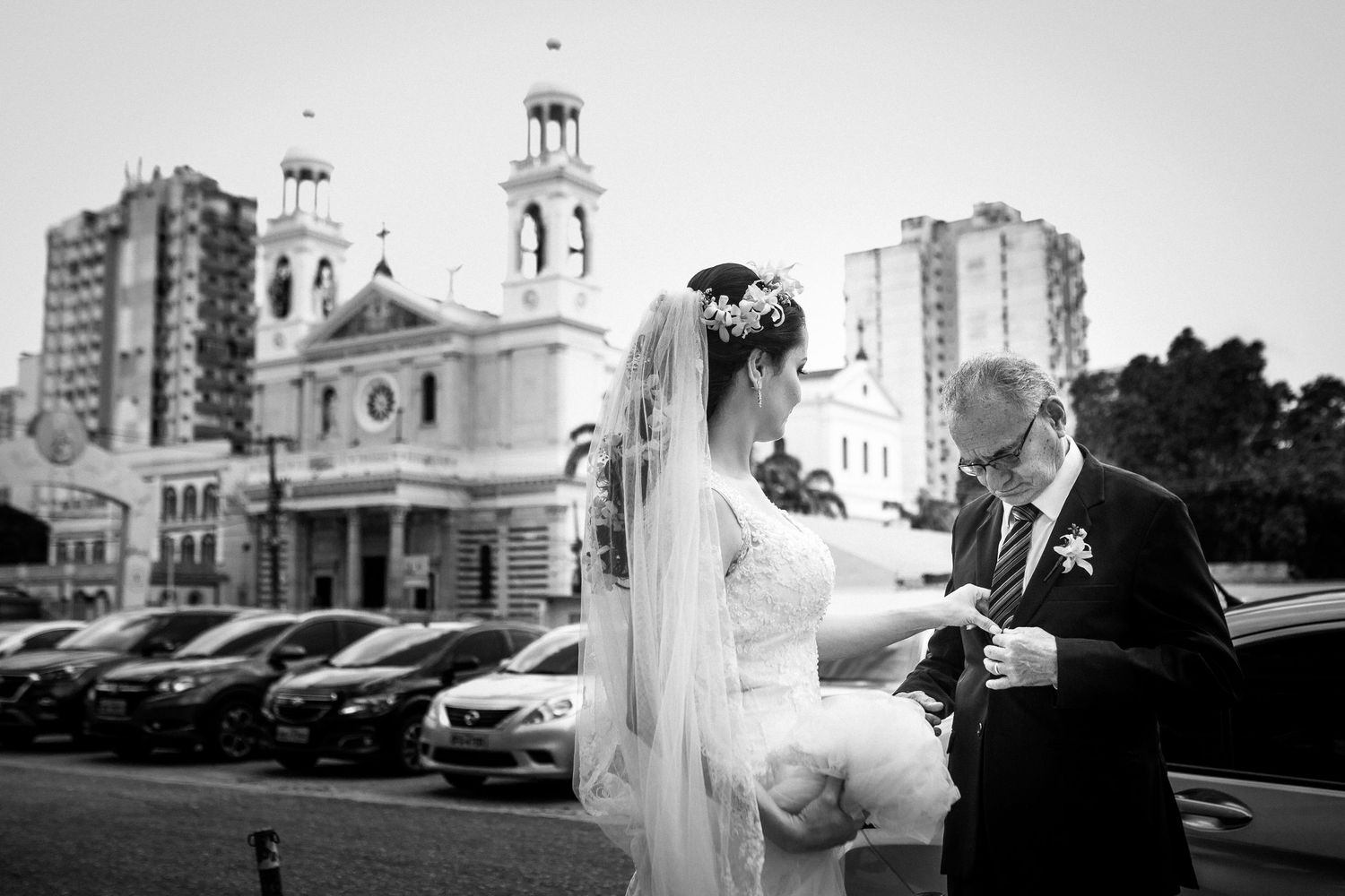 Black and white photo of a couple outside a church with parked cars in background.
