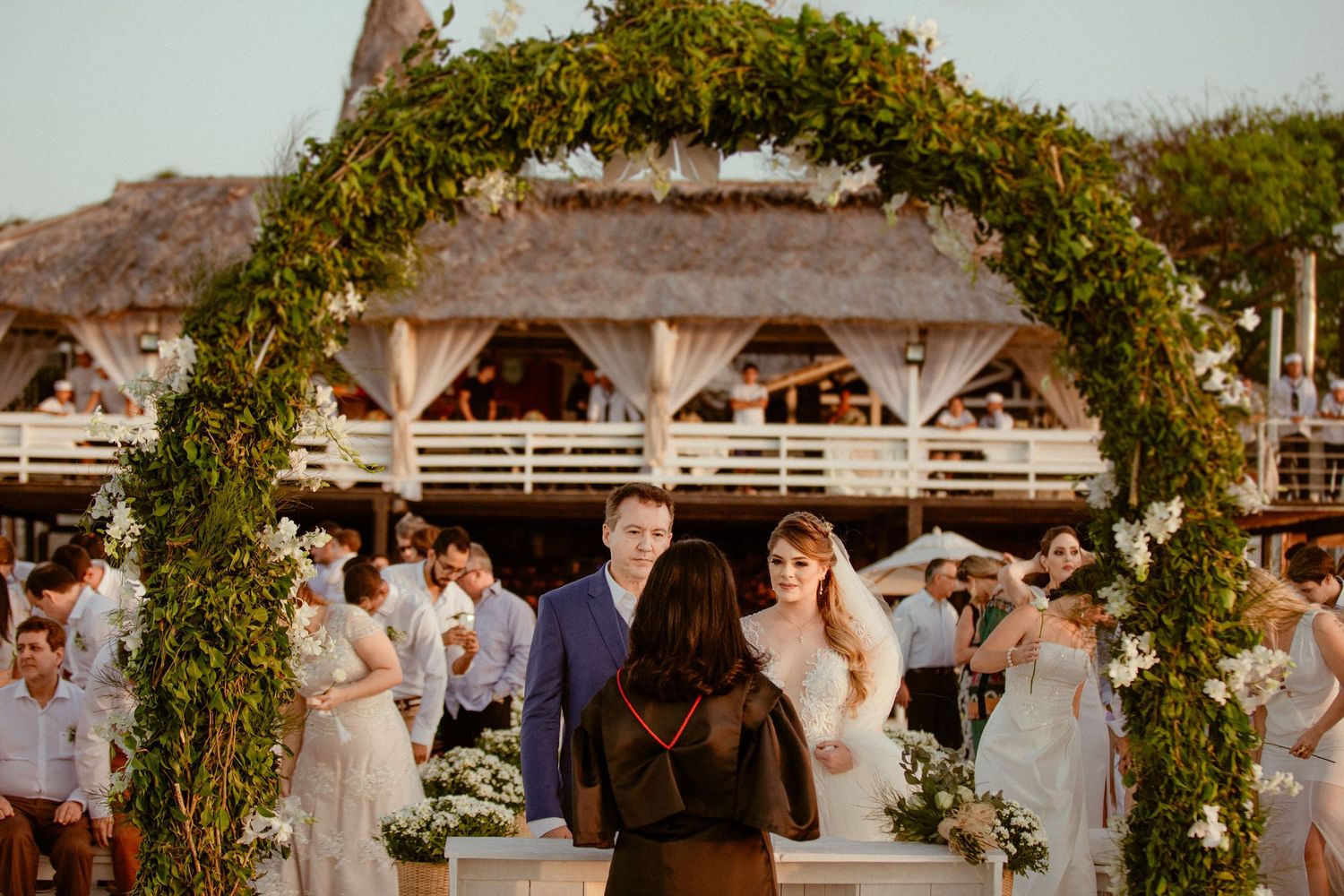 Wedding celebrations under an elegant floral and greenery arch with guests cheering at an outdoor venue during sunset.
