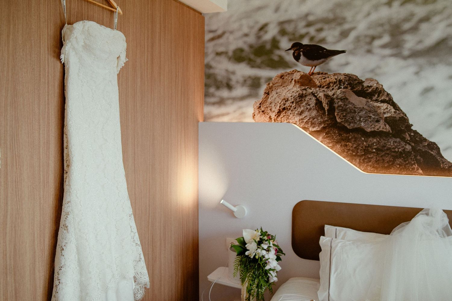 A white wedding dress hangs by a wooden wall next to a seagull perching on a branch and floral bouquet arrangement.