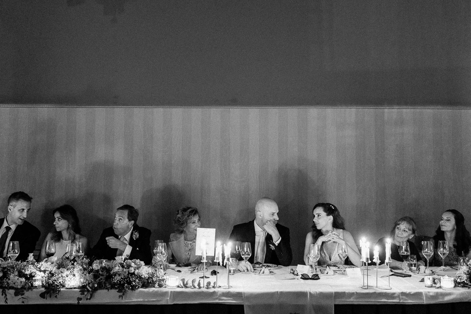 Black and white wedding reception photo of guests seated at a long head table with candles and floral arrangements.