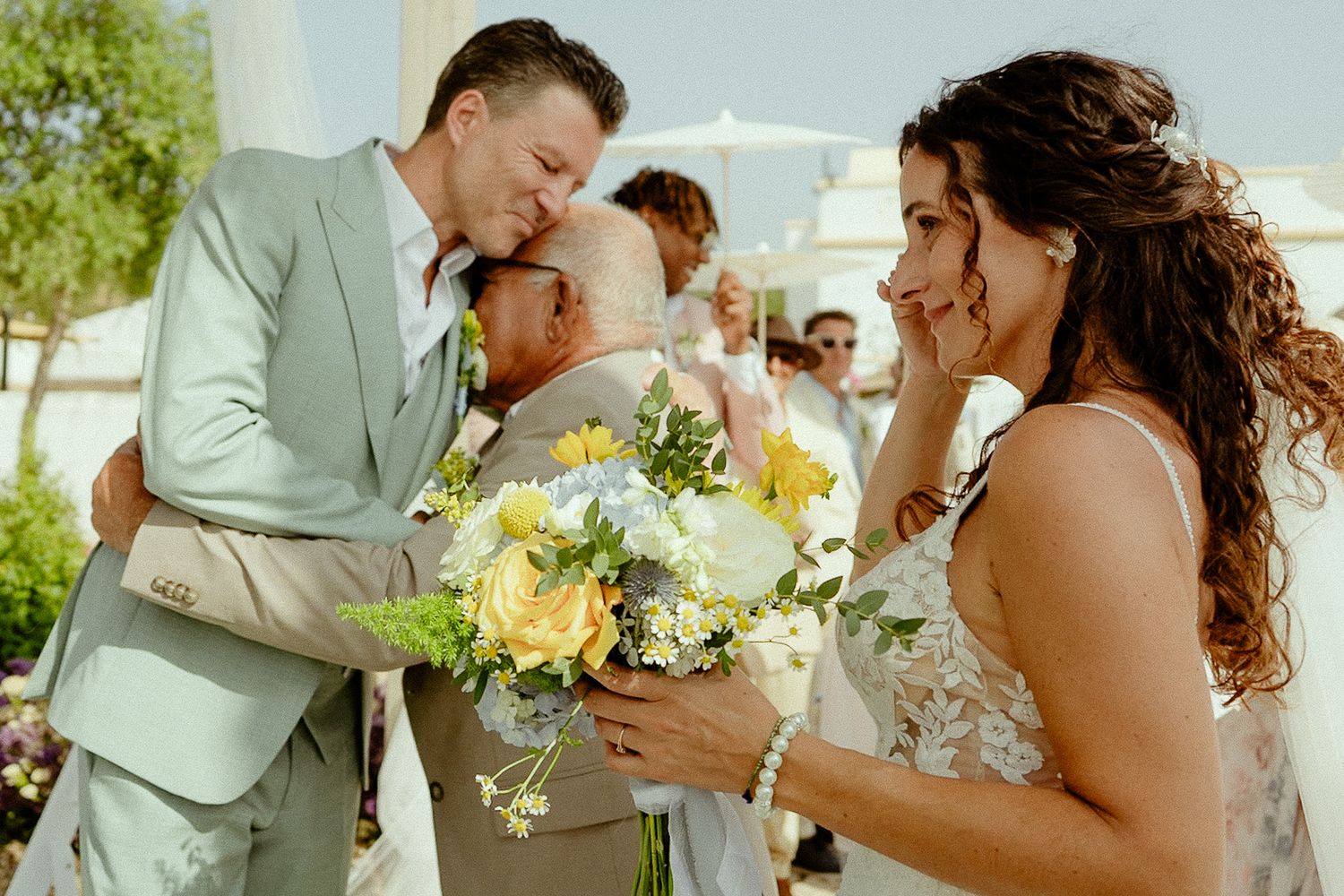 Bride and groom share emotional moment with guest at outdoor wedding, bride holding yellow and white bouquet.